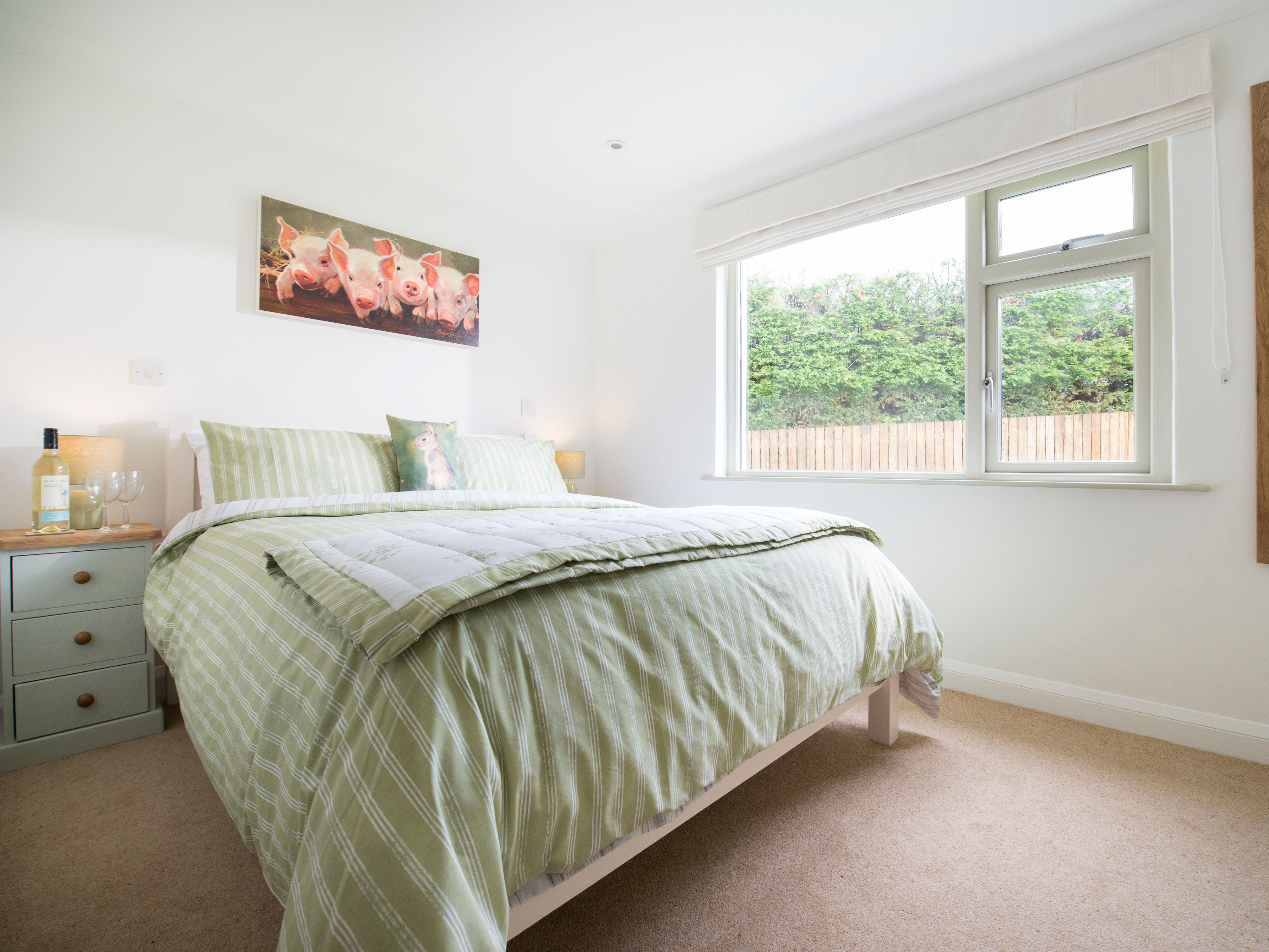 Bright bedroom with green striped bedding, a large window, a bedside table with a bottle of wine and glasses, a wall mirror, and a painting of piglets above the bed.