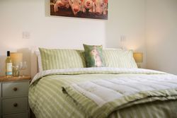 Cozy bedroom with green striped bedding, a rabbit pillow, bedside table with wine and glasses, and a painting of piglets on the wall.