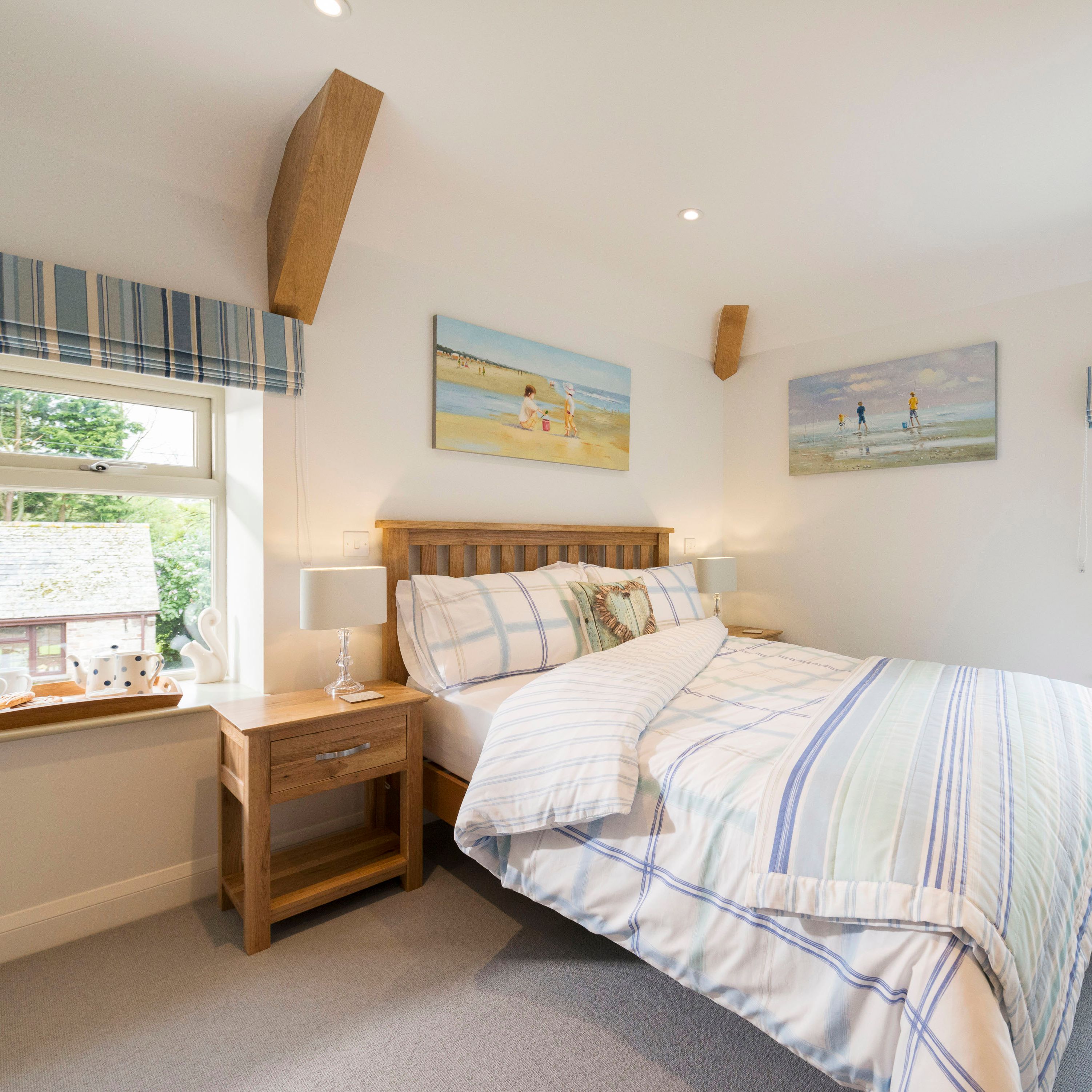 Bright, cozy bedroom with a wooden bed, plaid blue and white bedding, two windows with striped blue curtains, wooden nightstands with lamps, and beach-themed paintings on the walls.