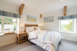 Bright, cozy bedroom with a wooden bed, plaid blue and white bedding, two windows with striped blue curtains, wooden nightstands with lamps, and beach-themed paintings on the walls.