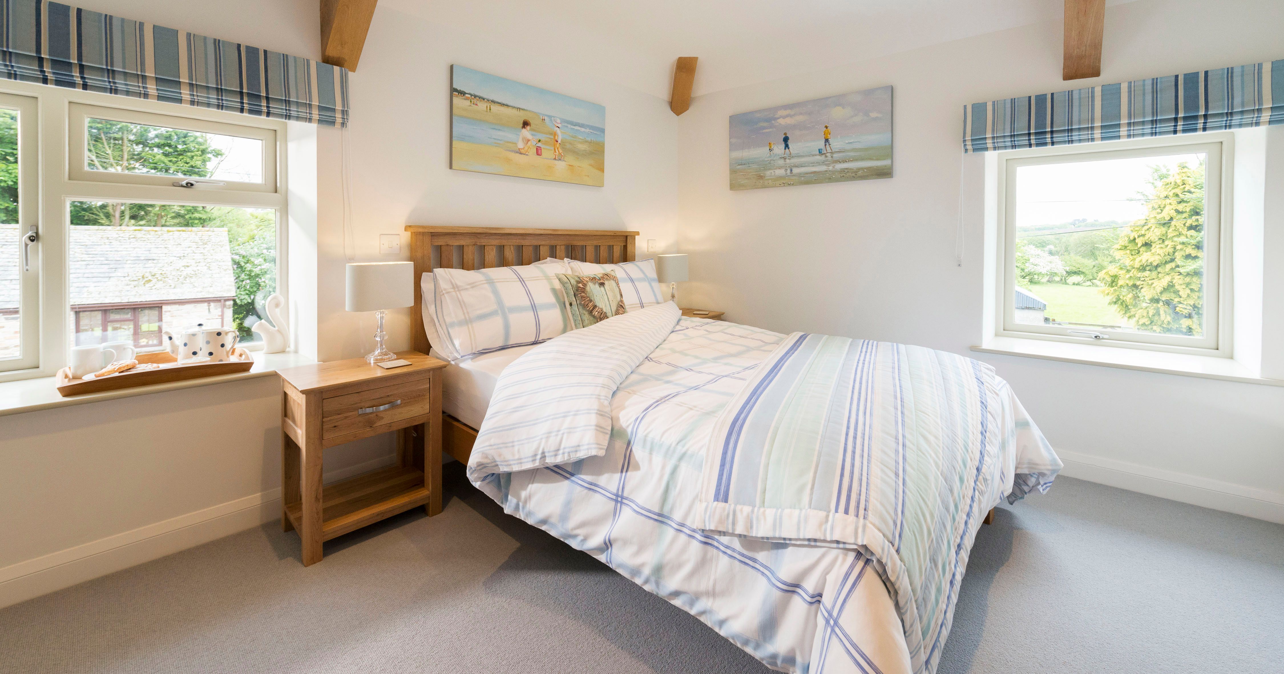 Bright, cozy bedroom with a wooden bed, plaid blue and white bedding, two windows with striped blue curtains, wooden nightstands with lamps, and beach-themed paintings on the walls.