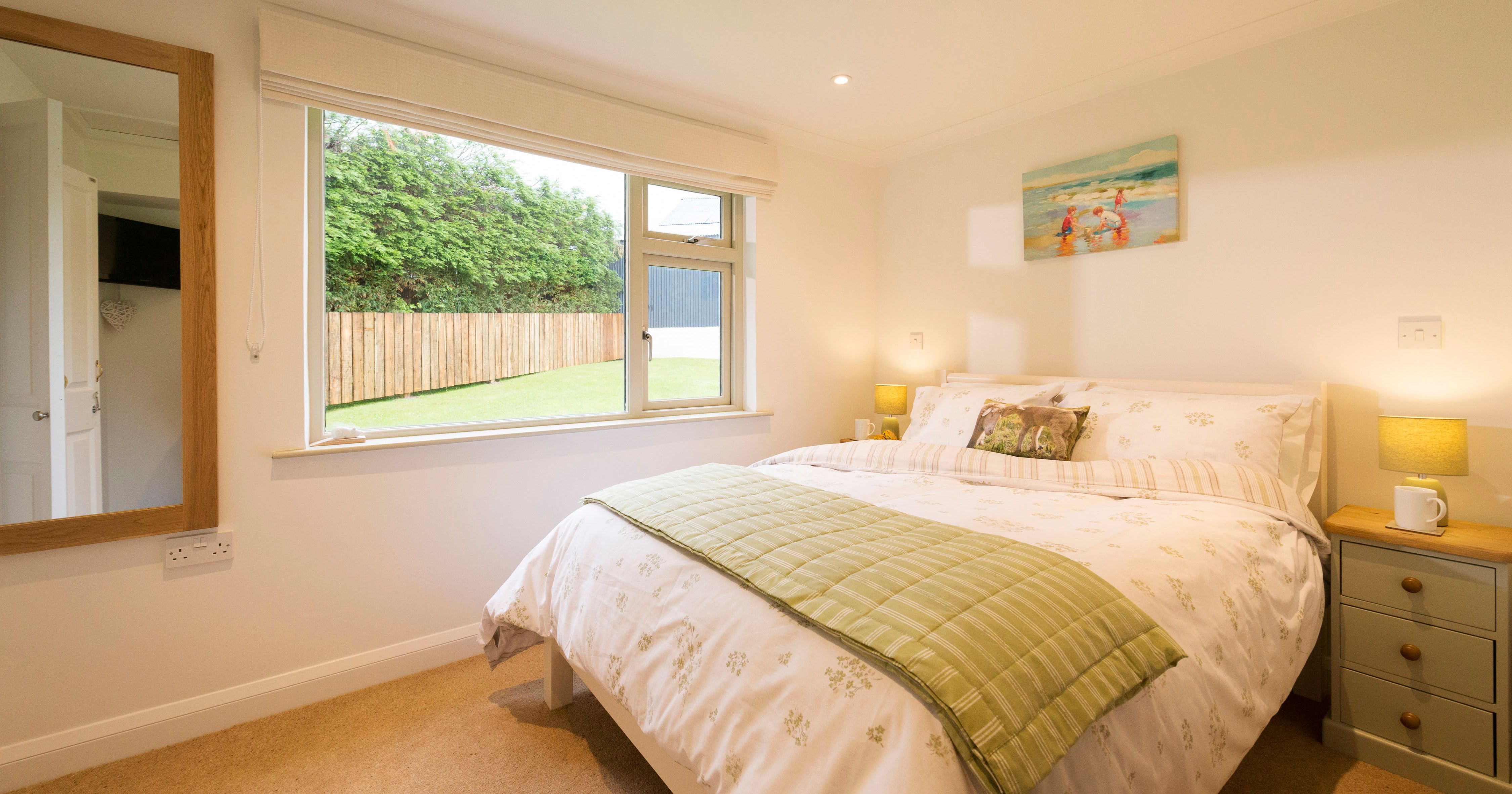 Bright and cozy bedroom with large window, double bed with floral bedding, green throw blanket, two bedside tables with lamps, a painting above the bed, and a mirror on the wall.