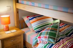 A cozy bedroom scene featuring a wooden bunk bed with colorful striped bedding, a wooden nightstand with an orange lamp, and two coasters.