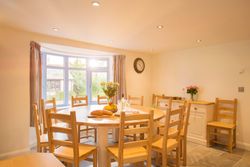 Bright dining room with a large wooden table surrounded by wooden chairs, a vase of flowers, bread, fruit, and wine on the table, large bay window with striped curtains, and a wall clock.