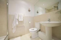 Modern bathroom with light beige tiles, white towel hanging on a heated towel rail, toilet with a book on the seat, pedestal sink, wall mirror, and shower area.