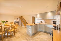 Modern open-plan kitchen and dining area with light grey cabinets, wooden countertops, tiled floor, round wooden dining table with chairs, and a staircase leading to the upper floor.
