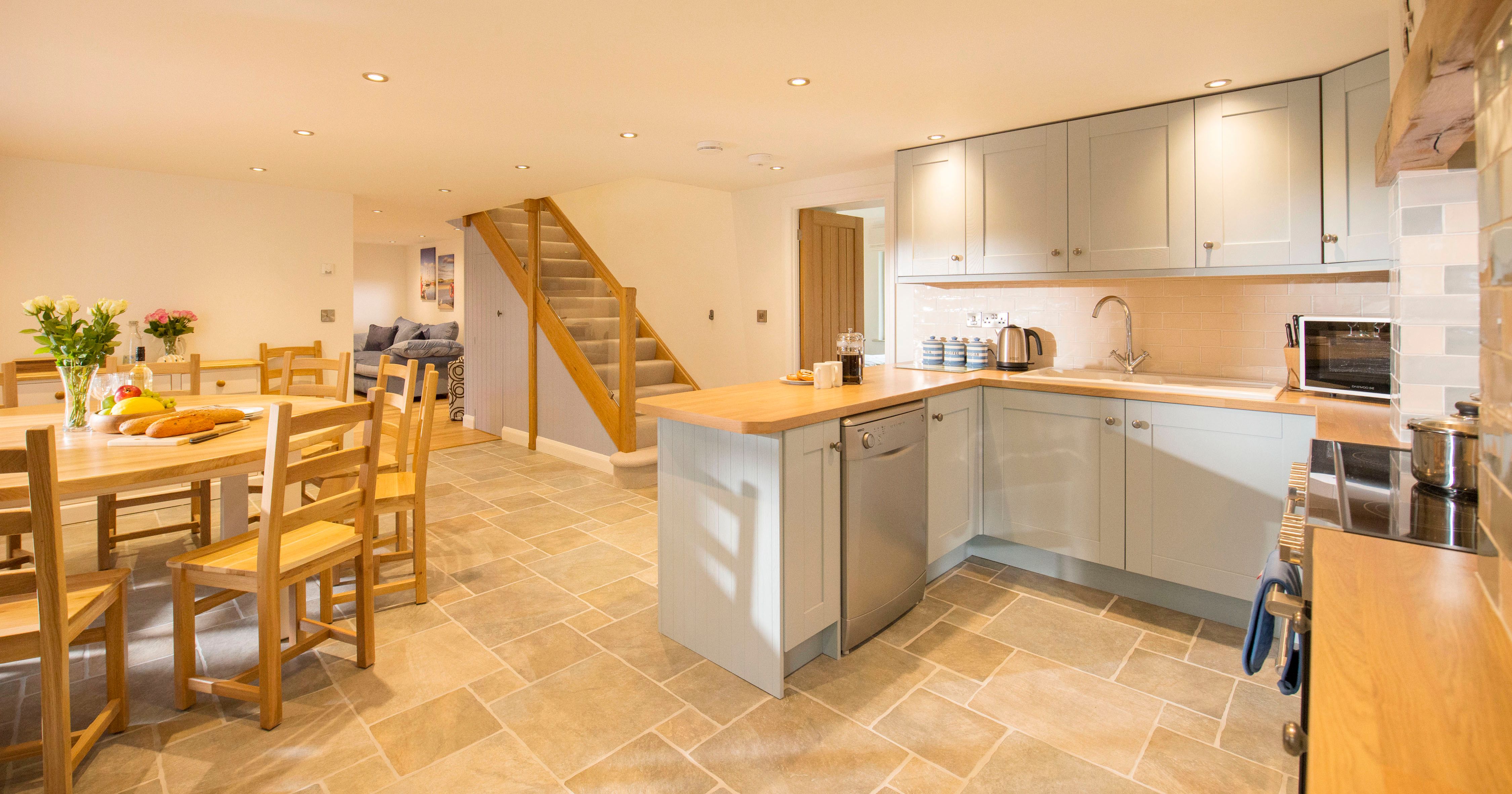 Modern open-plan kitchen and dining area with light grey cabinets, wooden countertops, tiled floor, round wooden dining table with chairs, and a staircase leading to the upper floor.