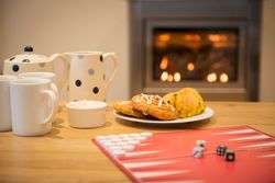 A cozy scene with a tea set, mugs, a plate of pastries, a bowl of sugar cubes, and a backgammon board on a wooden table in front of a lit fireplace.