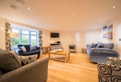 Spacious modern living room with wooden flooring, several sofas, a coffee table set for tea, patterned curtains, and a wall-mounted TV.