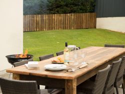 Outdoor wooden dining table set with plates, wine, glasses, and pastries on a patio next to a grill with fire, surrounded by wicker chairs in a grassy yard with a wooden fence.