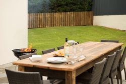 Outdoor wooden dining table set with plates, wine, glasses, and pastries on a patio next to a grill with fire, surrounded by wicker chairs in a grassy yard with a wooden fence.