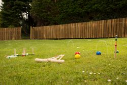 Croquet game set up on a grassy lawn with colorful balls, wooden mallets, and hoops; a wooden fence and trees are in the background.