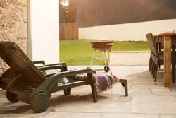 Outdoor patio area with green plastic lounge chairs, a blanket, a charcoal grill, wooden dining table with wicker chairs, and a green lawn enclosed by a wooden fence.