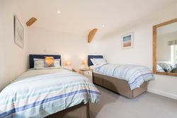 Bright and tidy twin bedroom with two single beds, striped blue and white bedding, bedside table with lamps, wall art, large mirror, and a window letting in natural light.