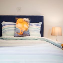 A neatly made bed with striped bedding, featuring a pillow with a picture of a yellow duckling. Next to the bed is a nightstand with a lamp and a plate of pastries.