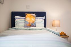 A neatly made bed with striped bedding, featuring a pillow with a picture of a yellow duckling. Next to the bed is a nightstand with a lamp and a plate of pastries.