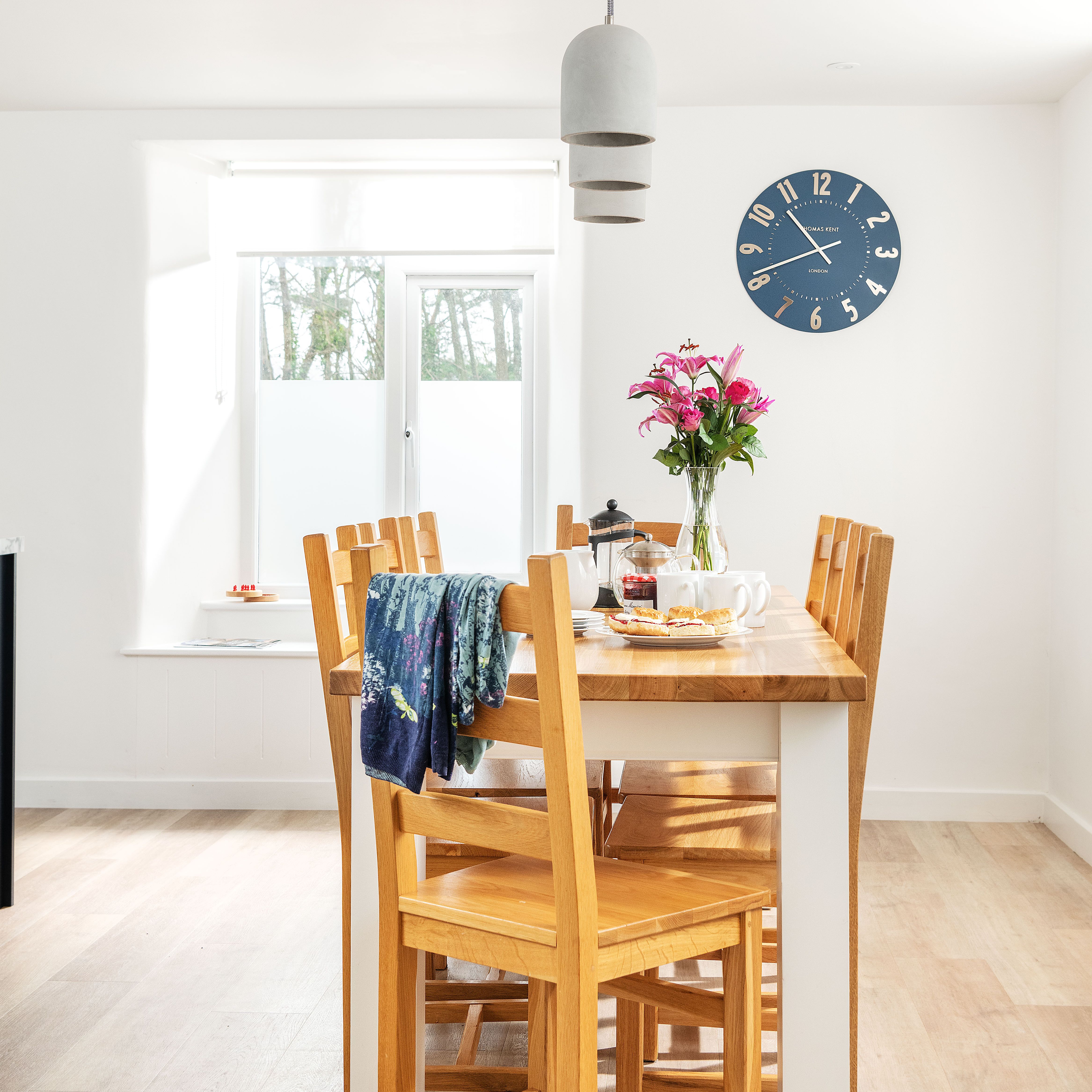 Bright dining room with wooden table and chairs, a vase of pink flowers, breakfast items, blue wall clock, pendant light, and large window.