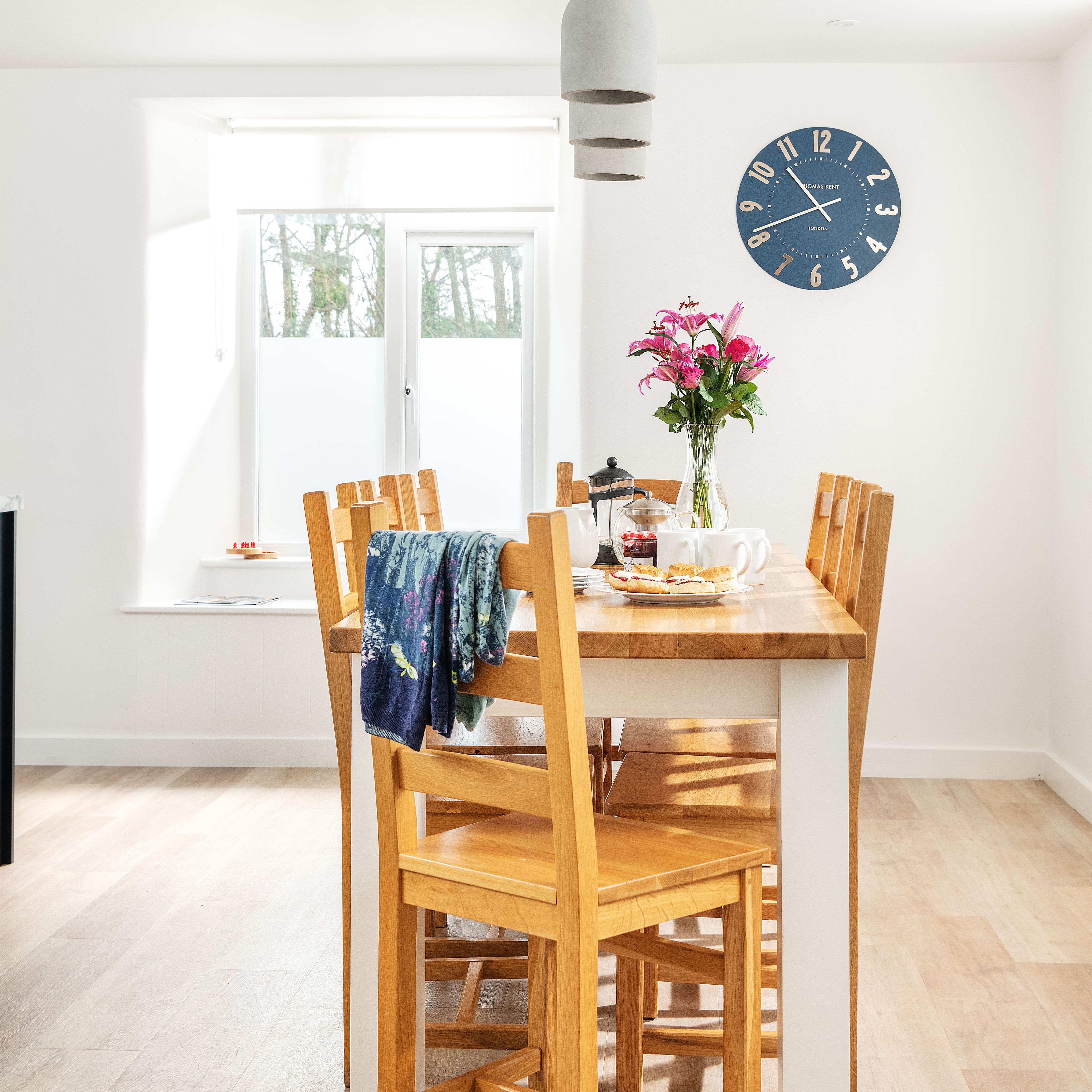 Bright dining room with wooden table and chairs, a vase of pink flowers, breakfast items, blue wall clock, pendant light, and large window.