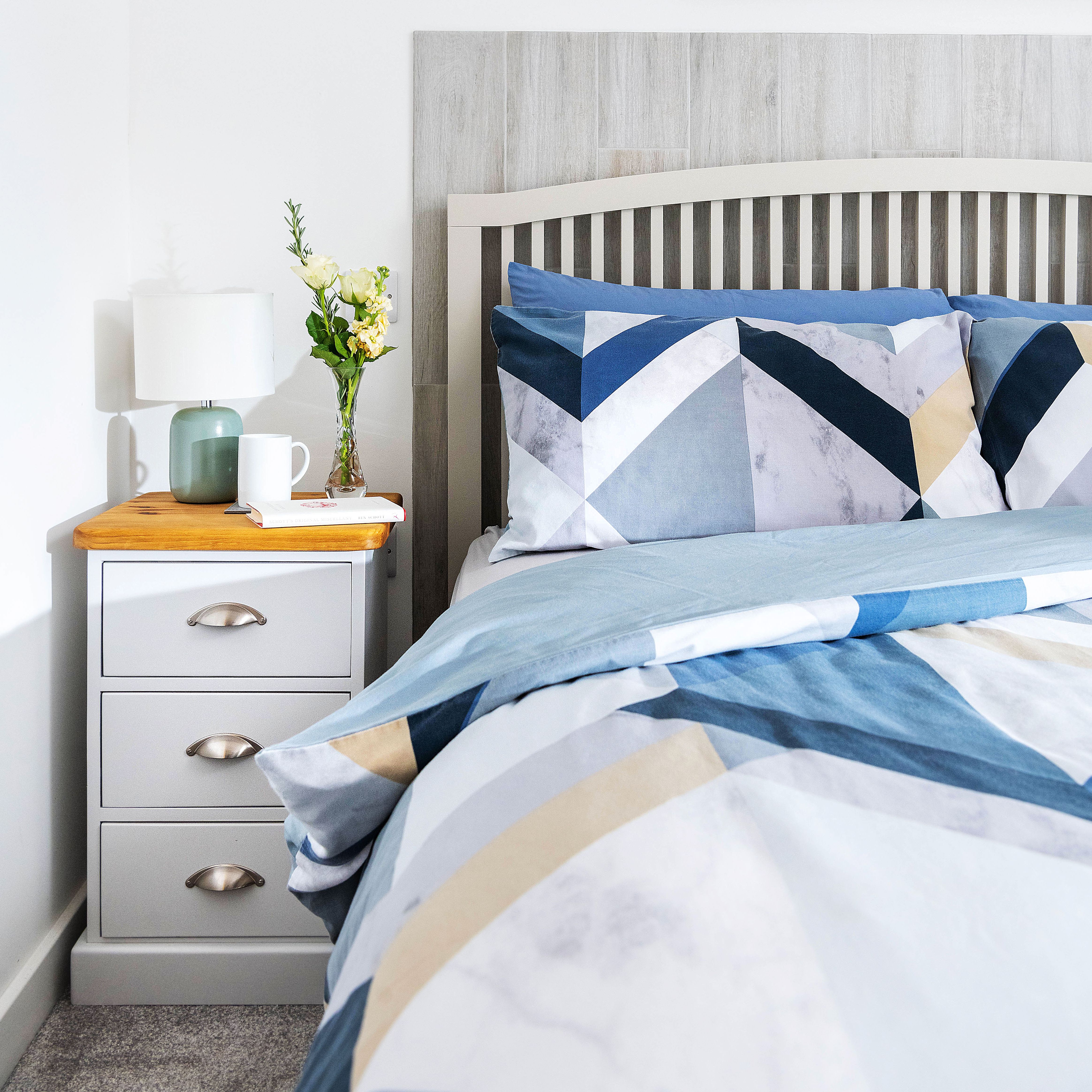 Modern bedroom with geometric bedding, a wooden headboard, a nightstand with a lamp, vase of flowers, mug, and a book, and a coastal painting on the wall.