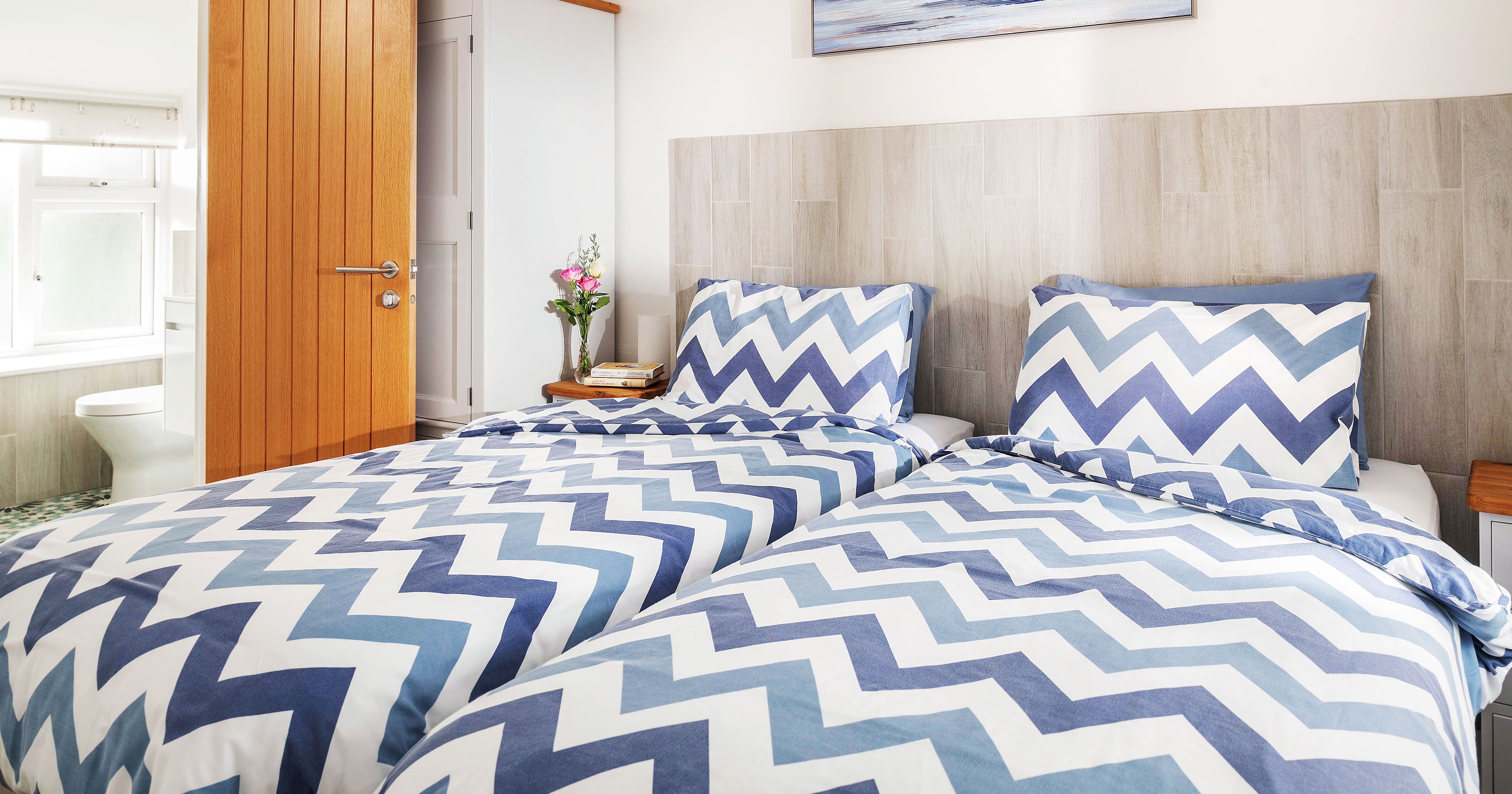 Bright bedroom with two single beds featuring blue and white chevron bedding, a wooden door leading to a bathroom, a nightstand with flowers and books, and modern decor.