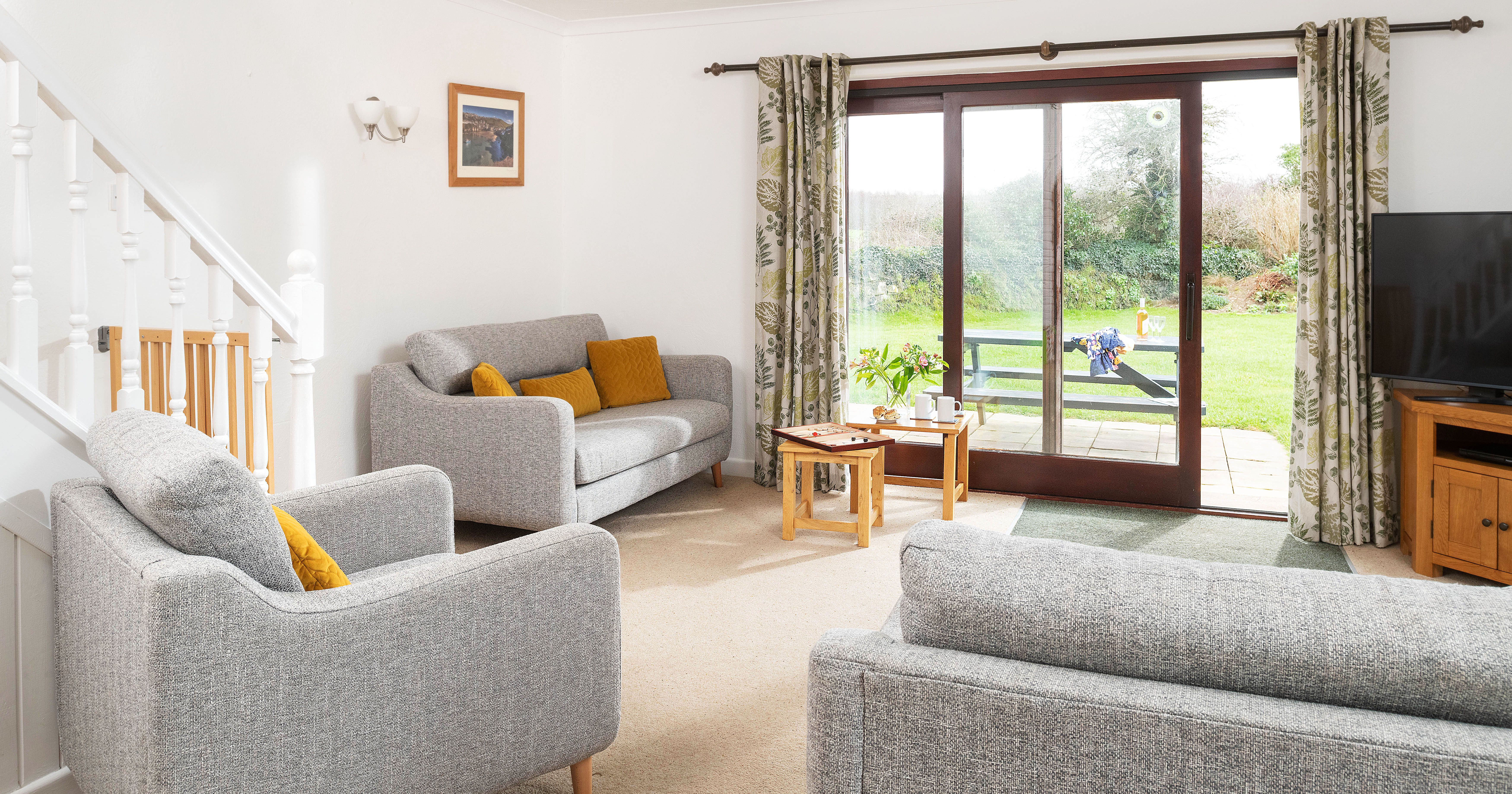 Bright living room with gray sofas, mustard yellow cushions, coffee table, large sliding glass doors opening to a green garden, and a TV on a wooden stand.