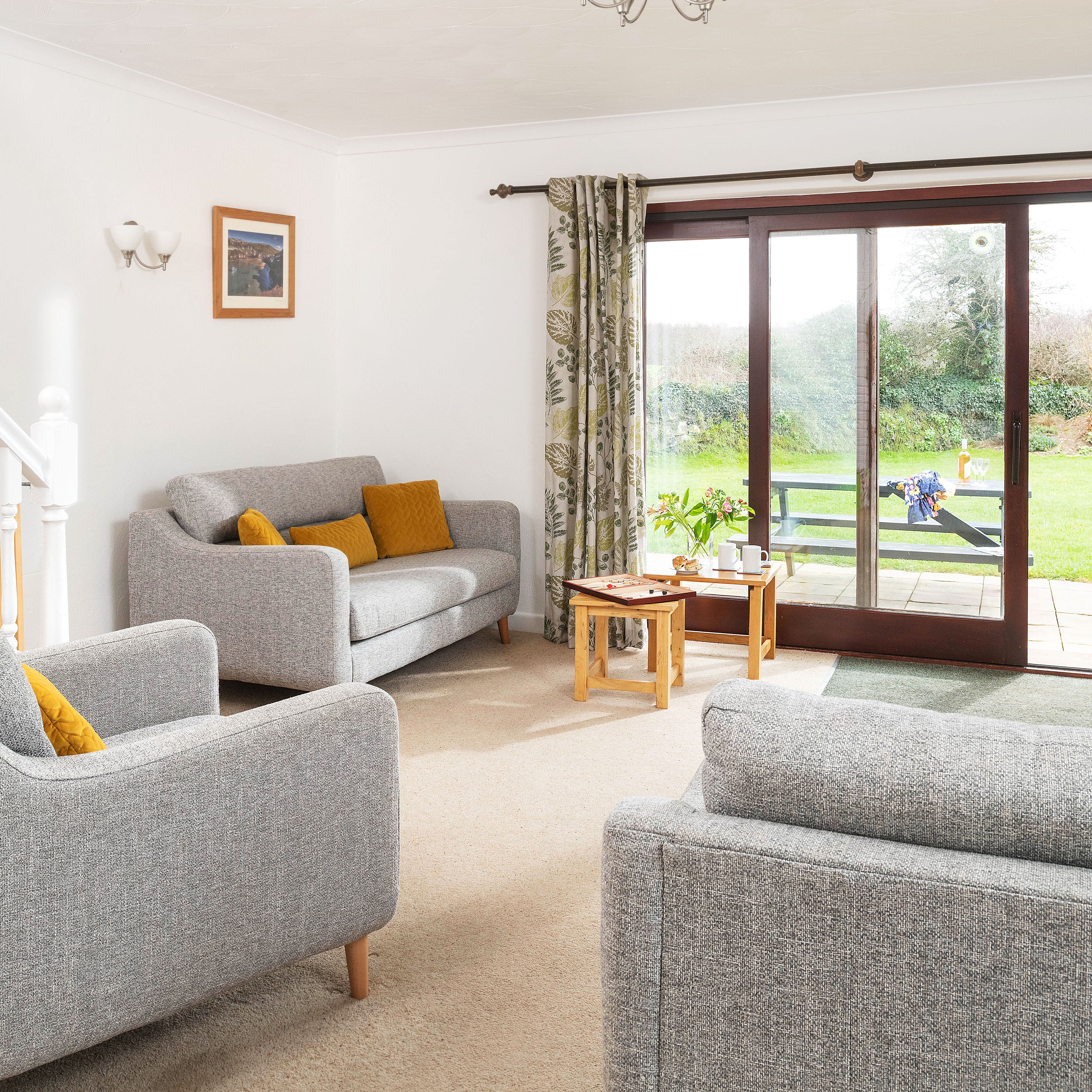 Bright living room with gray sofas, mustard yellow cushions, coffee table, large sliding glass doors opening to a green garden, and a TV on a wooden stand.