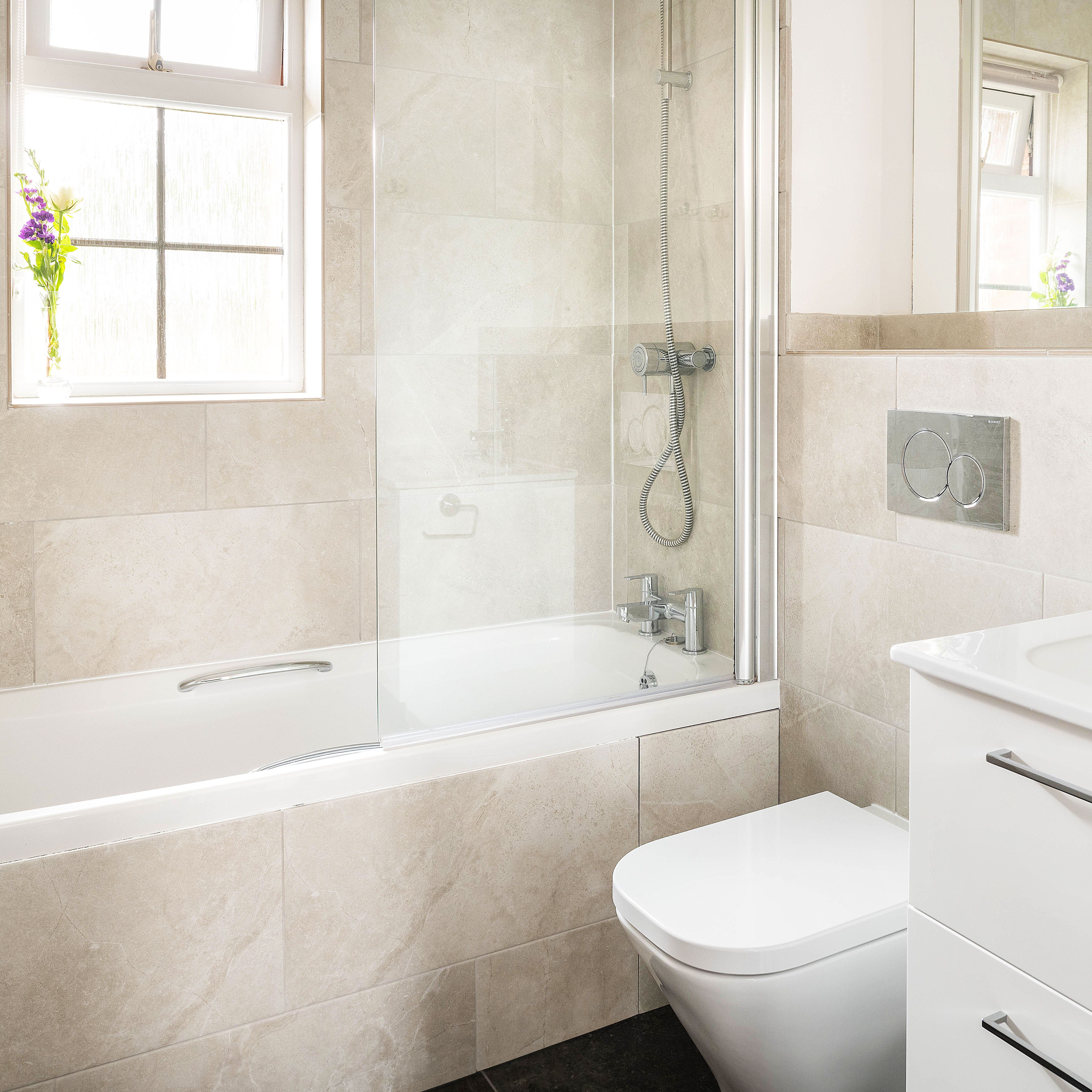 Modern bathroom with a bathtub, shower, wall-mounted toilet, white vanity with sink, large mirror, and a small vase with purple flowers on the window sill.