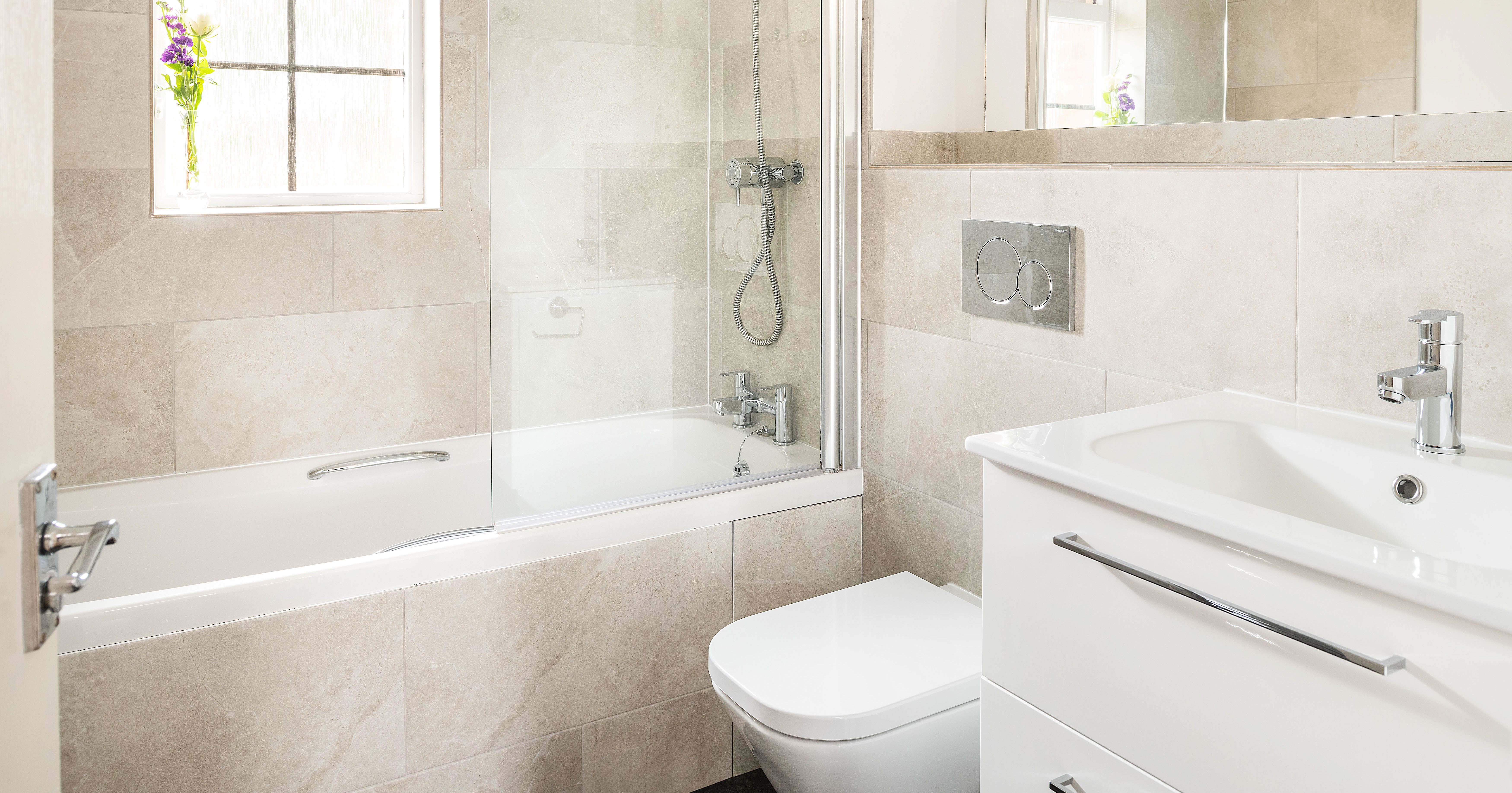 Modern bathroom with a bathtub, shower, wall-mounted toilet, white vanity with sink, large mirror, and a small vase with purple flowers on the window sill.