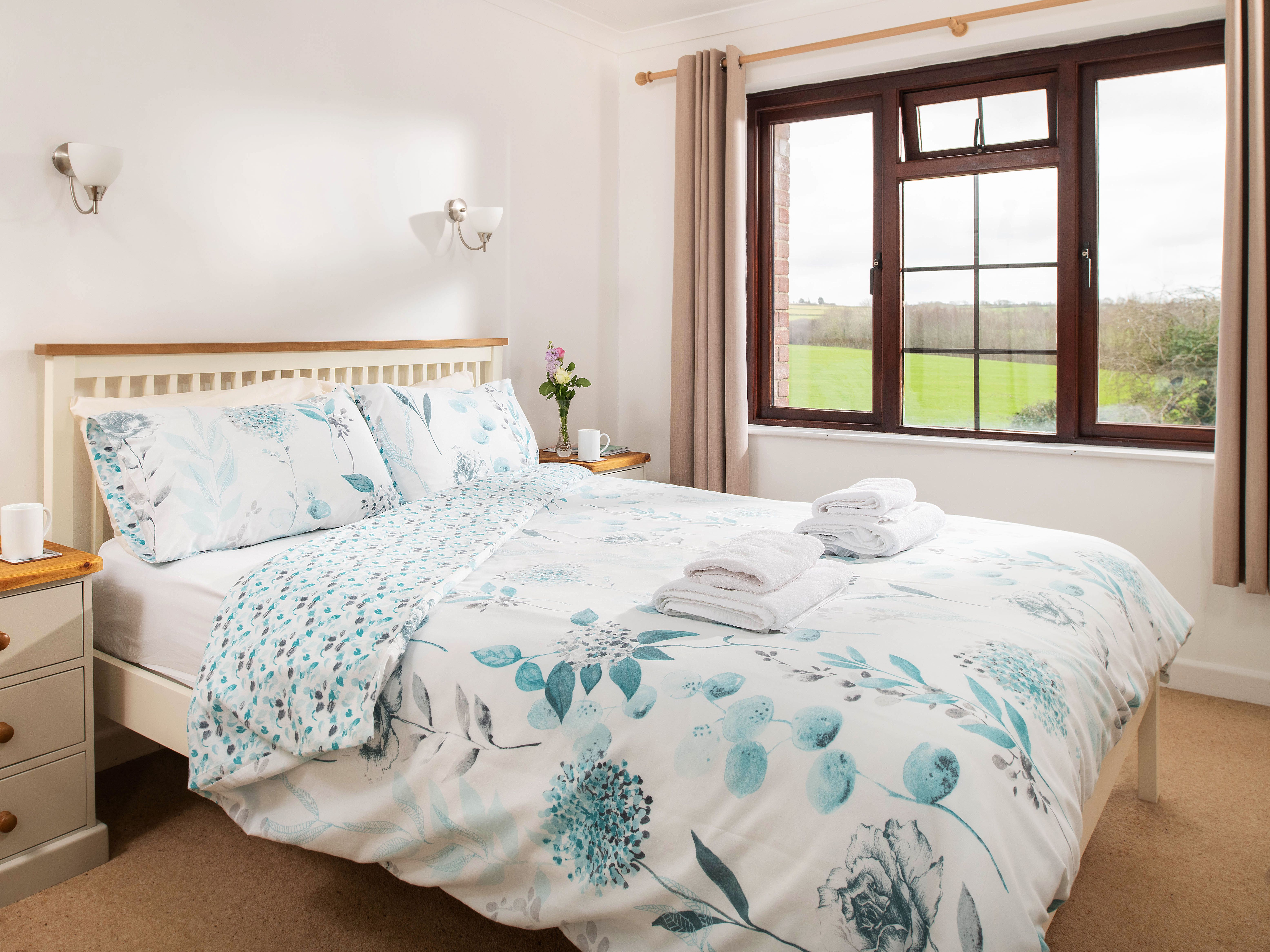 Bright and cozy bedroom with a double bed, floral bedding, bedside tables with cups and books, a window with curtains, and a view of green fields outside.