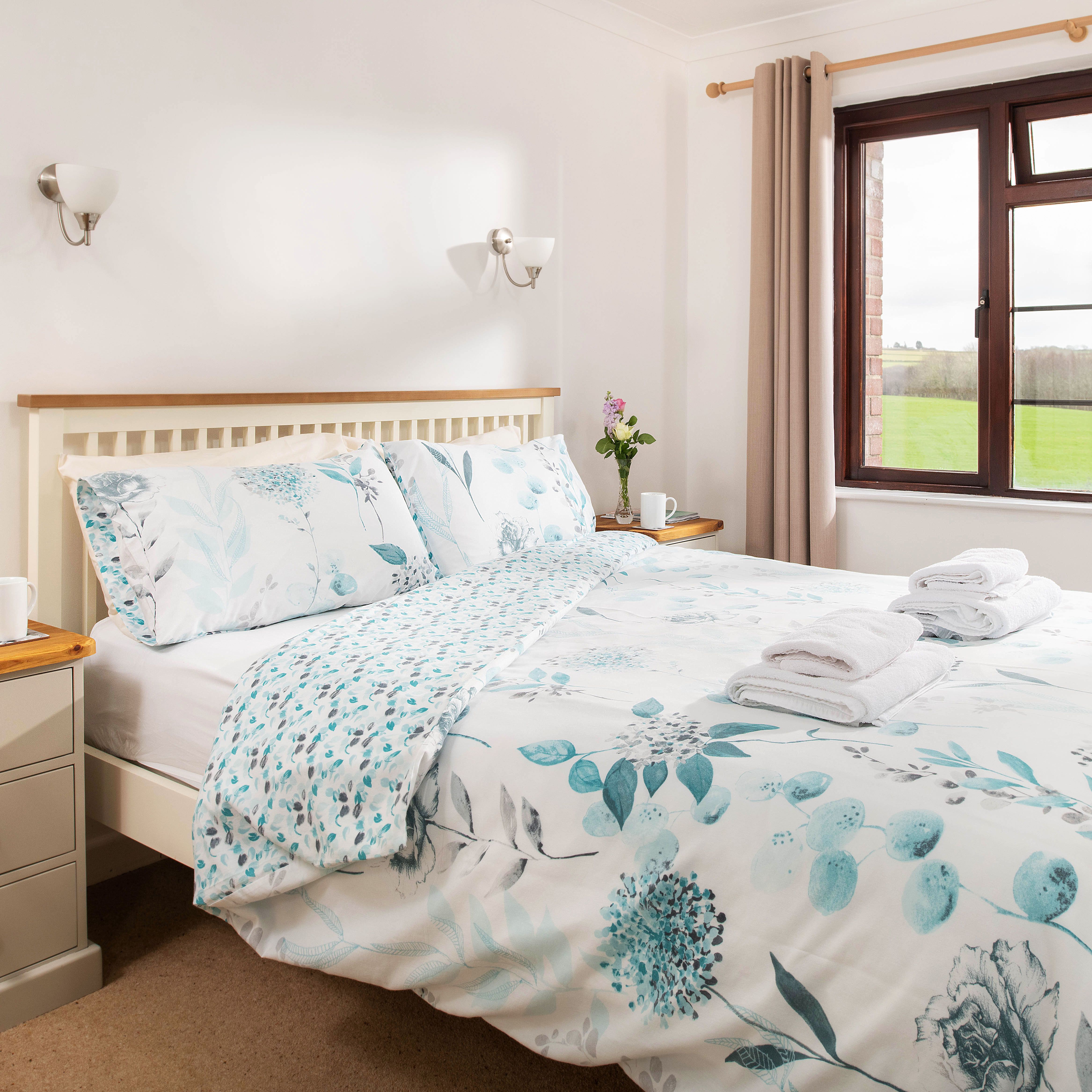 Bright and cozy bedroom with a double bed, floral bedding, bedside tables with cups and books, a window with curtains, and a view of green fields outside.