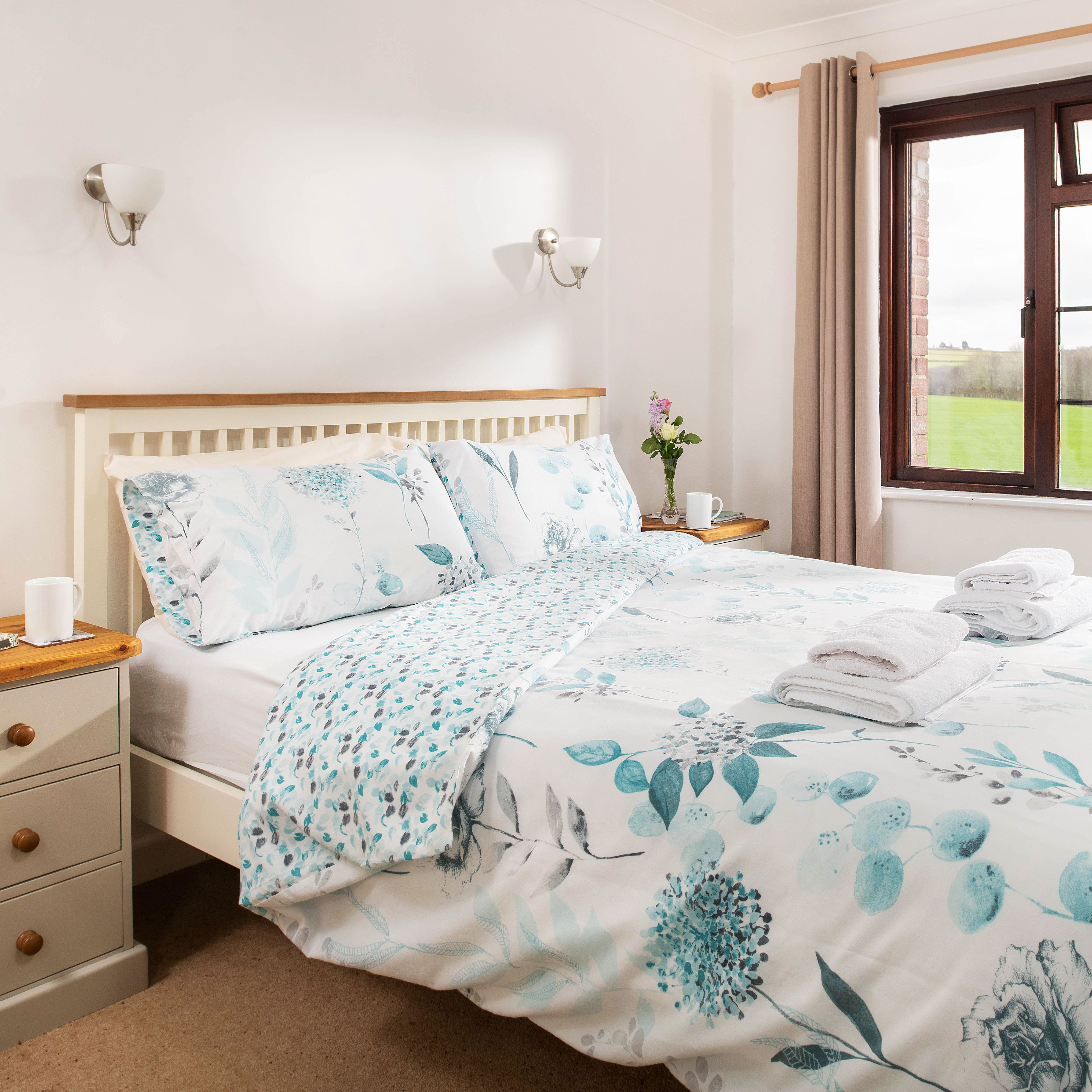 Bright and cozy bedroom with a double bed, floral bedding, bedside tables with cups and books, a window with curtains, and a view of green fields outside.