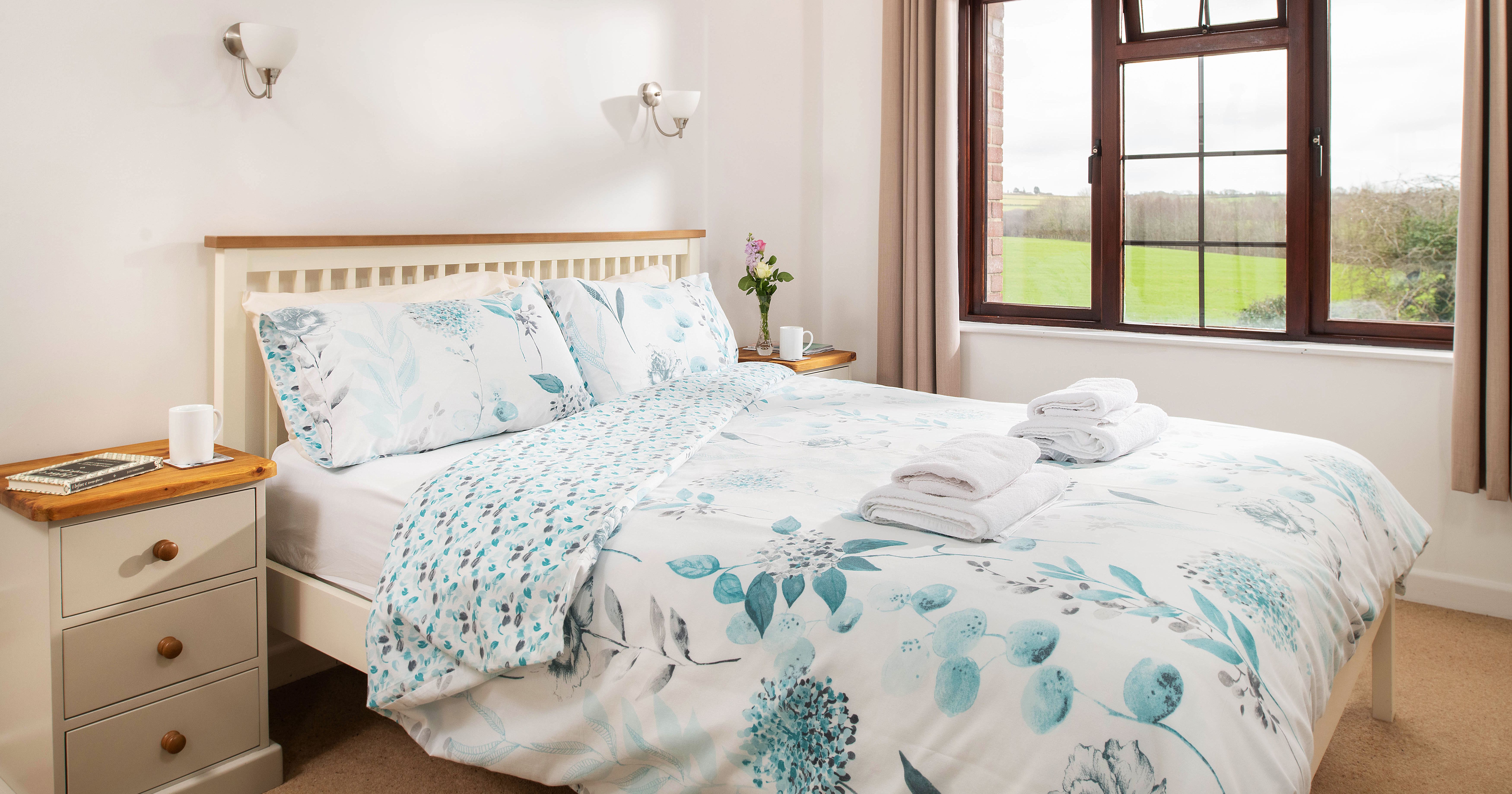 Bright and cozy bedroom with a double bed, floral bedding, bedside tables with cups and books, a window with curtains, and a view of green fields outside.