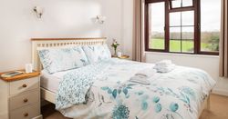 Bright and cozy bedroom with a double bed, floral bedding, bedside tables with cups and books, a window with curtains, and a view of green fields outside.