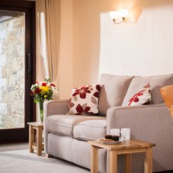Cozy living room with a beige sofa, floral cushions, small wooden tables with snacks and mugs, a vase of flowers, and a wooden dining table with a tea set.