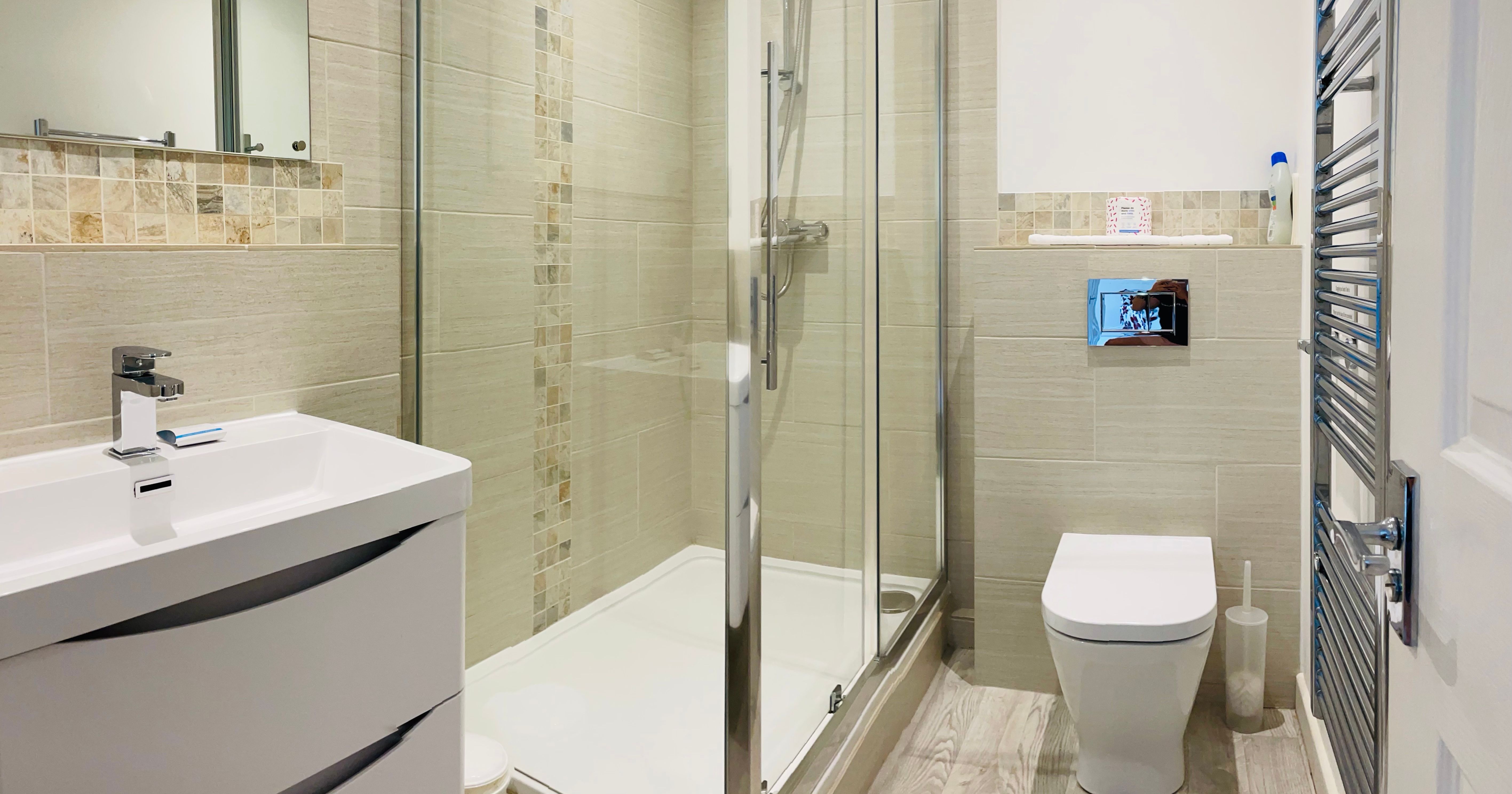Modern bathroom with glass shower enclosure, white sink cabinet, wall-mounted toilet, tiled walls and floor, and chrome towel radiator.