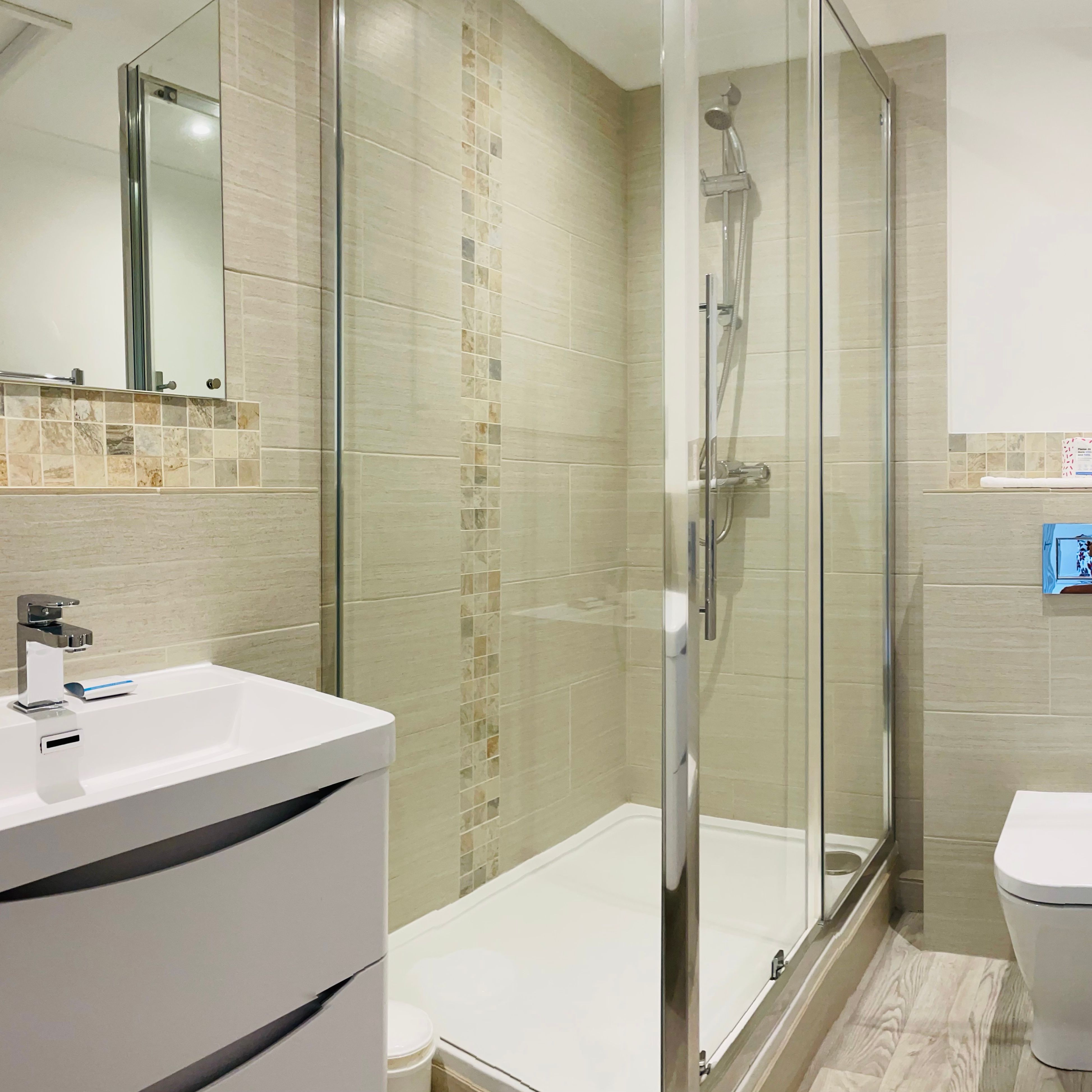 Modern bathroom with glass shower enclosure, white sink cabinet, wall-mounted toilet, tiled walls and floor, and chrome towel radiator.