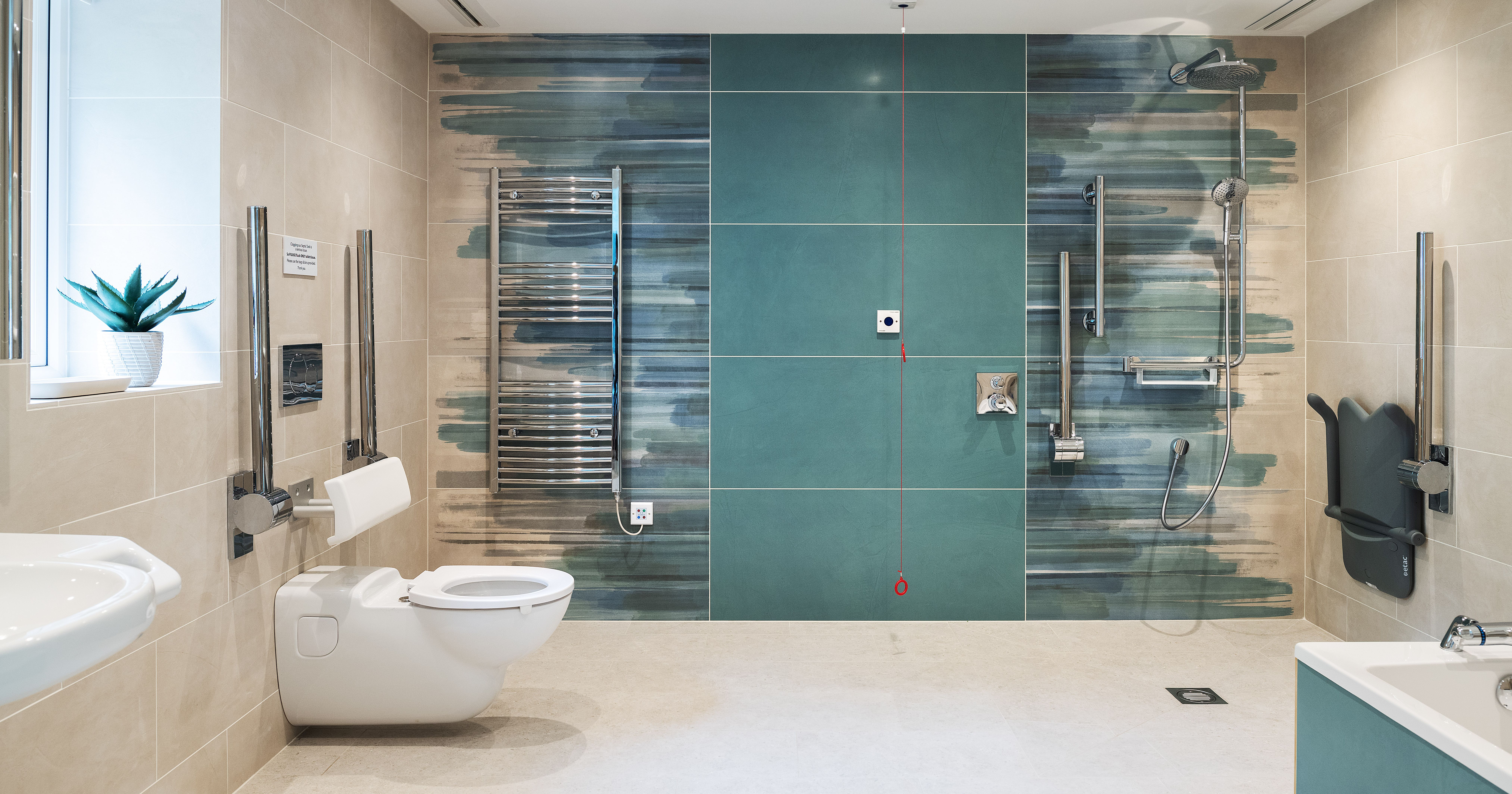 Modern accessible bathroom with wall-mounted toilet, grab bars, open shower area with seating and handrails, heated towel rack, emergency pull cord, and stylish teal and beige tile design.