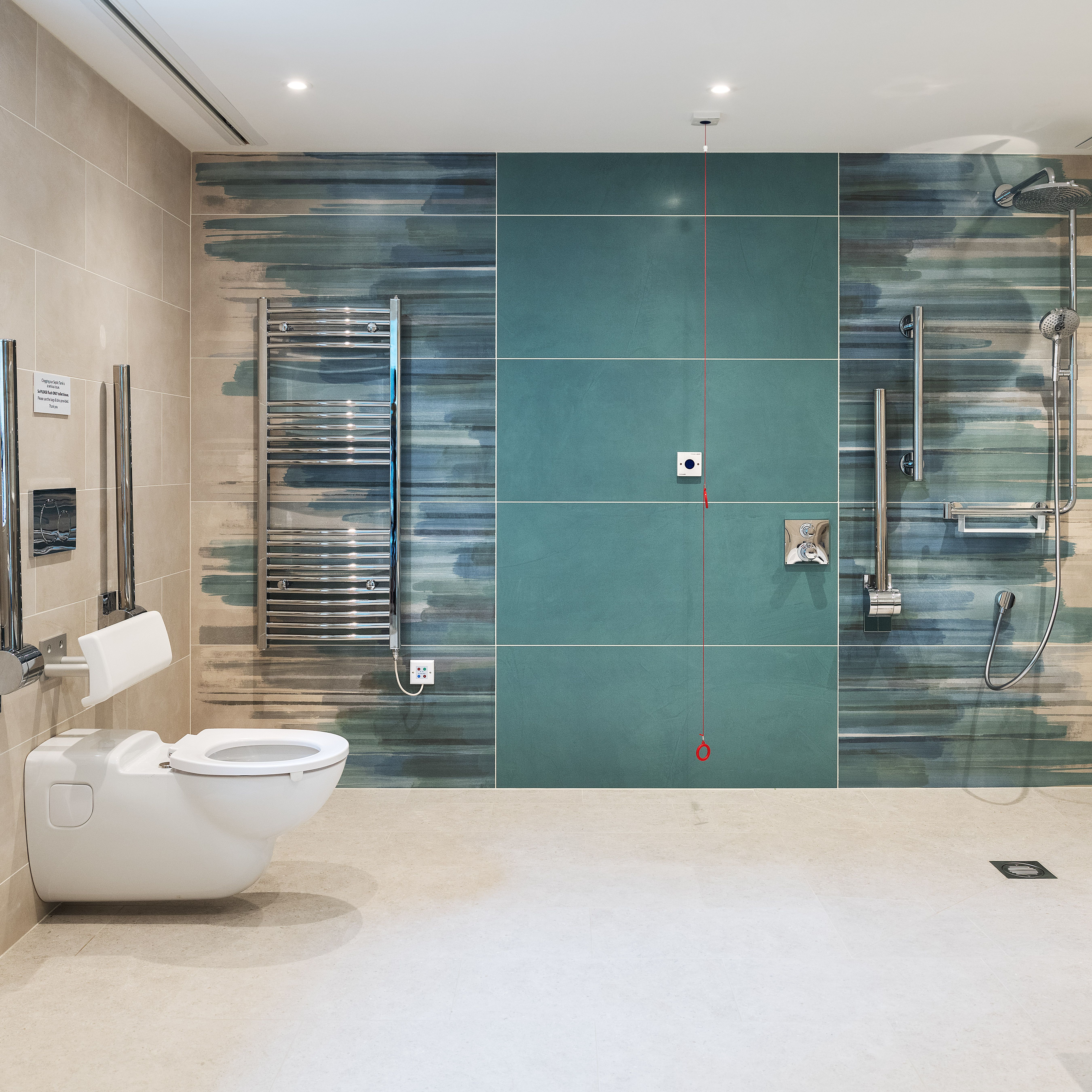 Modern accessible bathroom with wall-mounted toilet, grab bars, open shower area with seating and handrails, heated towel rack, emergency pull cord, and stylish teal and beige tile design.