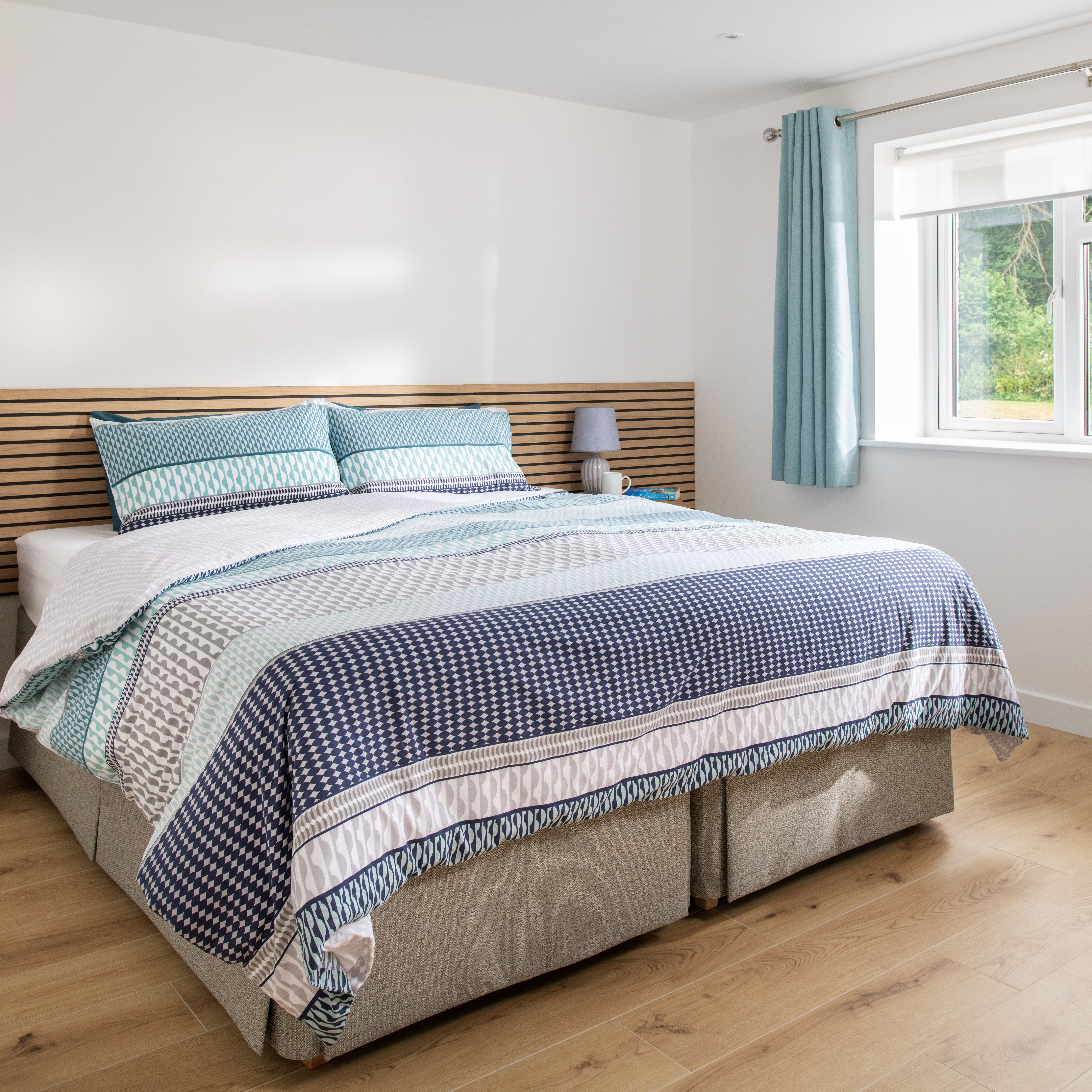 Bright modern bedroom with a large bed, patterned bedding in shades of blue and white, wooden floors, bedside tables with lamps, and a window with blue curtains letting in natural light.