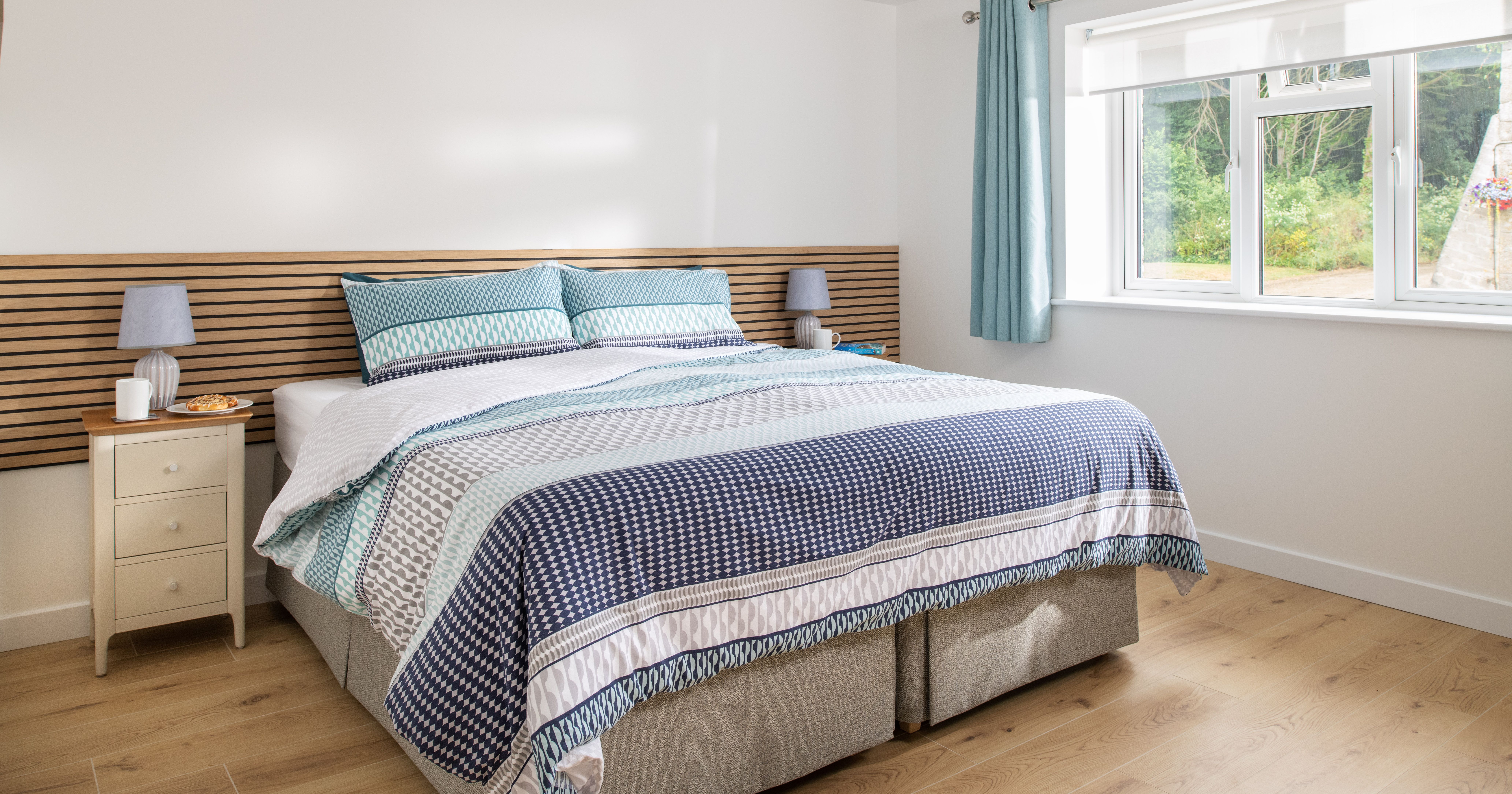 Bright modern bedroom with a large bed, patterned bedding in shades of blue and white, wooden floors, bedside tables with lamps, and a window with blue curtains letting in natural light.