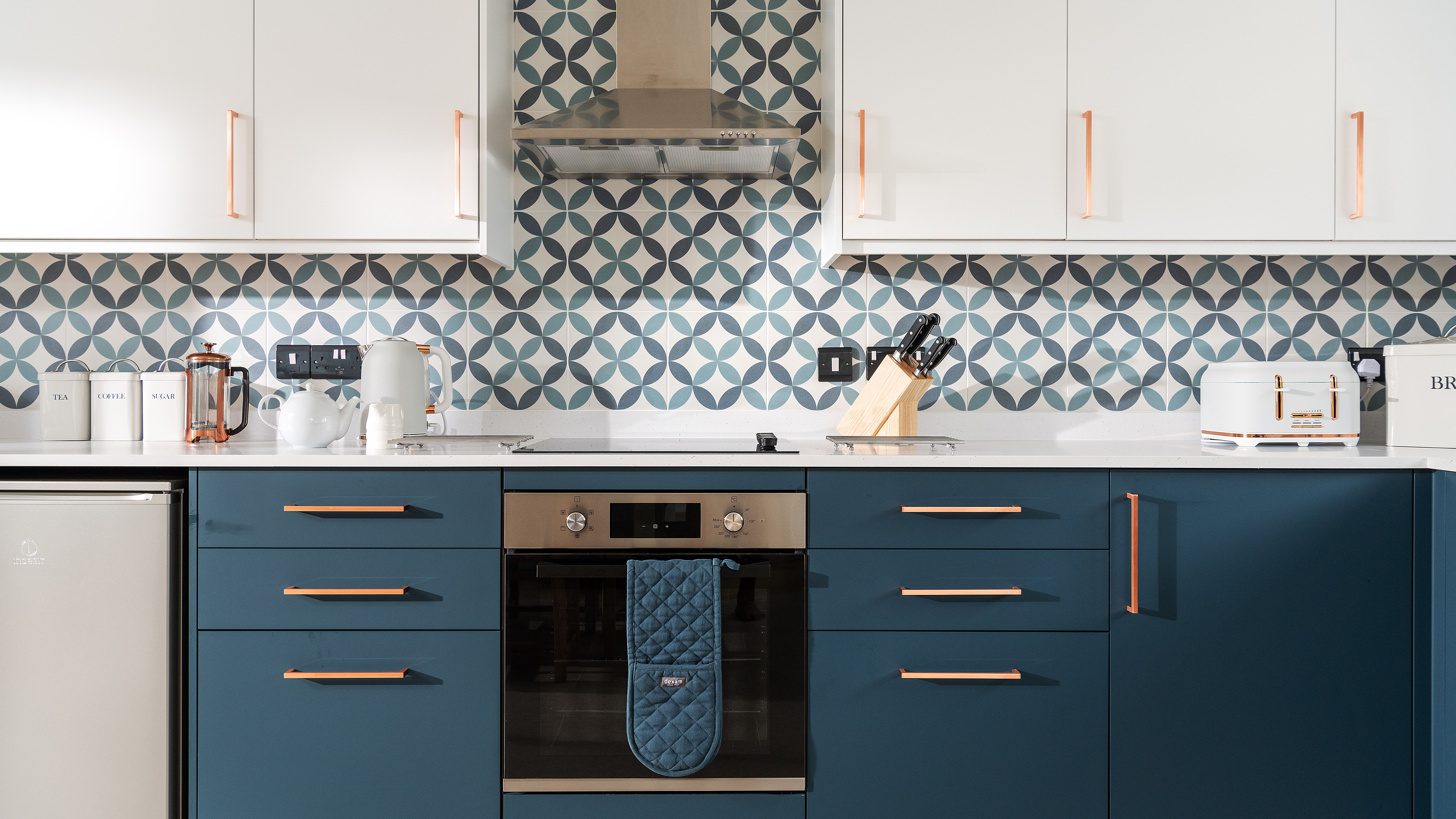 Modern kitchen with blue cabinets, patterned tile backsplash, white upper cabinets, stainless steel oven, and kitchen appliances on the countertop.