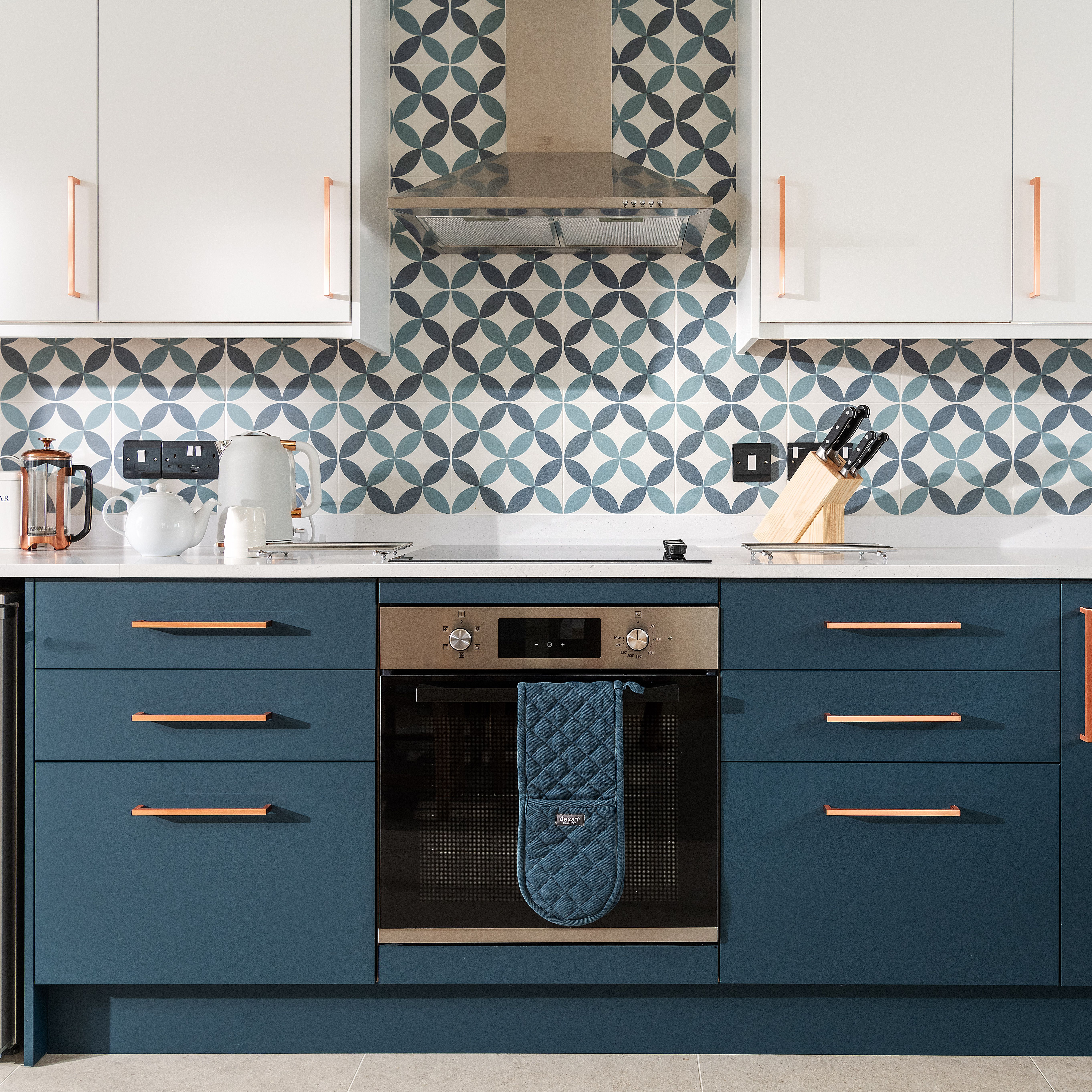 Modern kitchen with blue cabinets, patterned tile backsplash, white upper cabinets, stainless steel oven, and kitchen appliances on the countertop.
