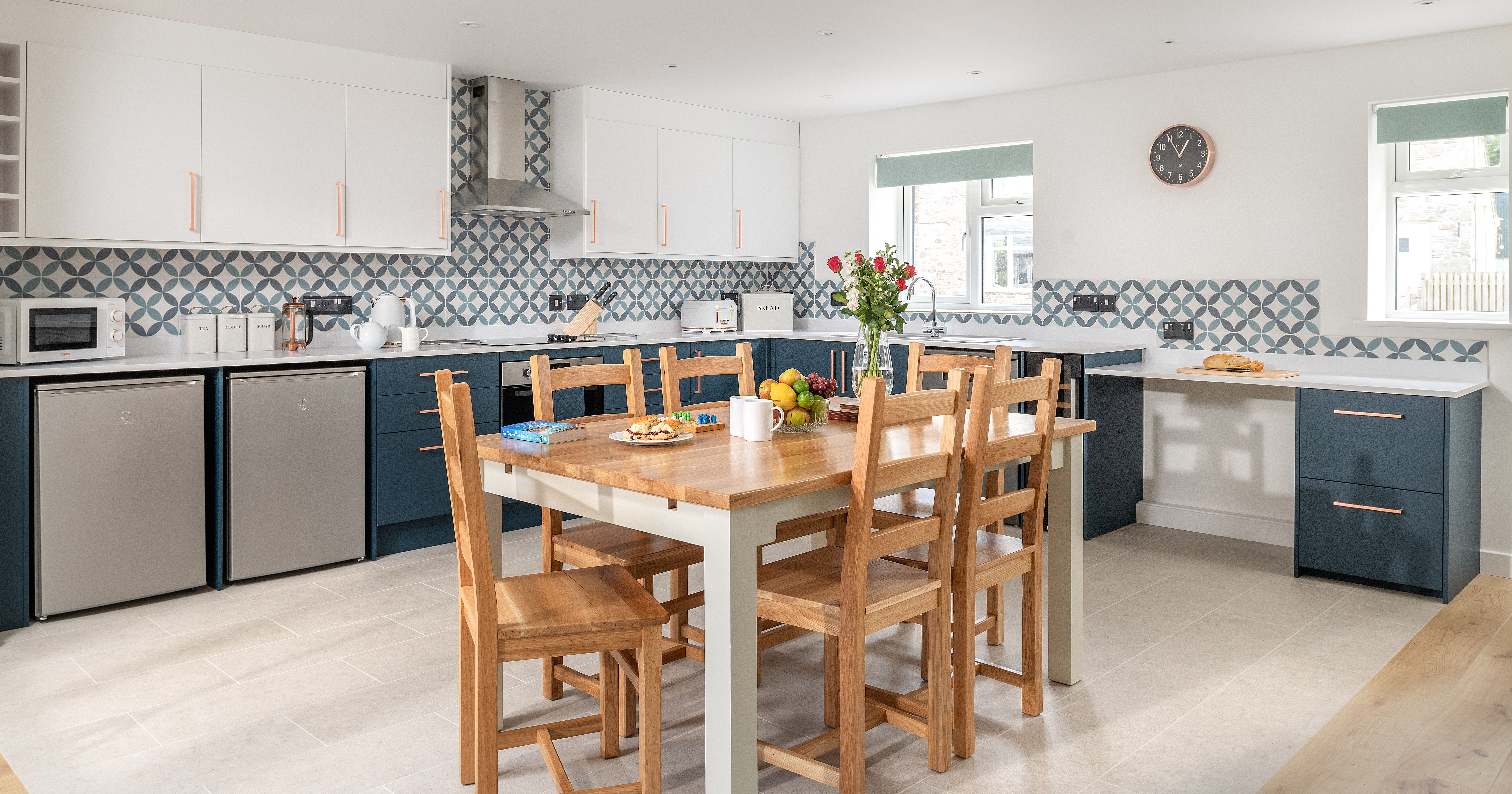 Modern kitchen with patterned backsplash, wooden dining table and chairs, blue and white cabinets, stainless steel appliances, and a vase of flowers on the table.