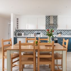 Modern kitchen with blue and white cabinets, patterned backsplash, wooden dining table with six chairs, vase of flowers and a bowl of fruit on the table, and stainless steel appliances.