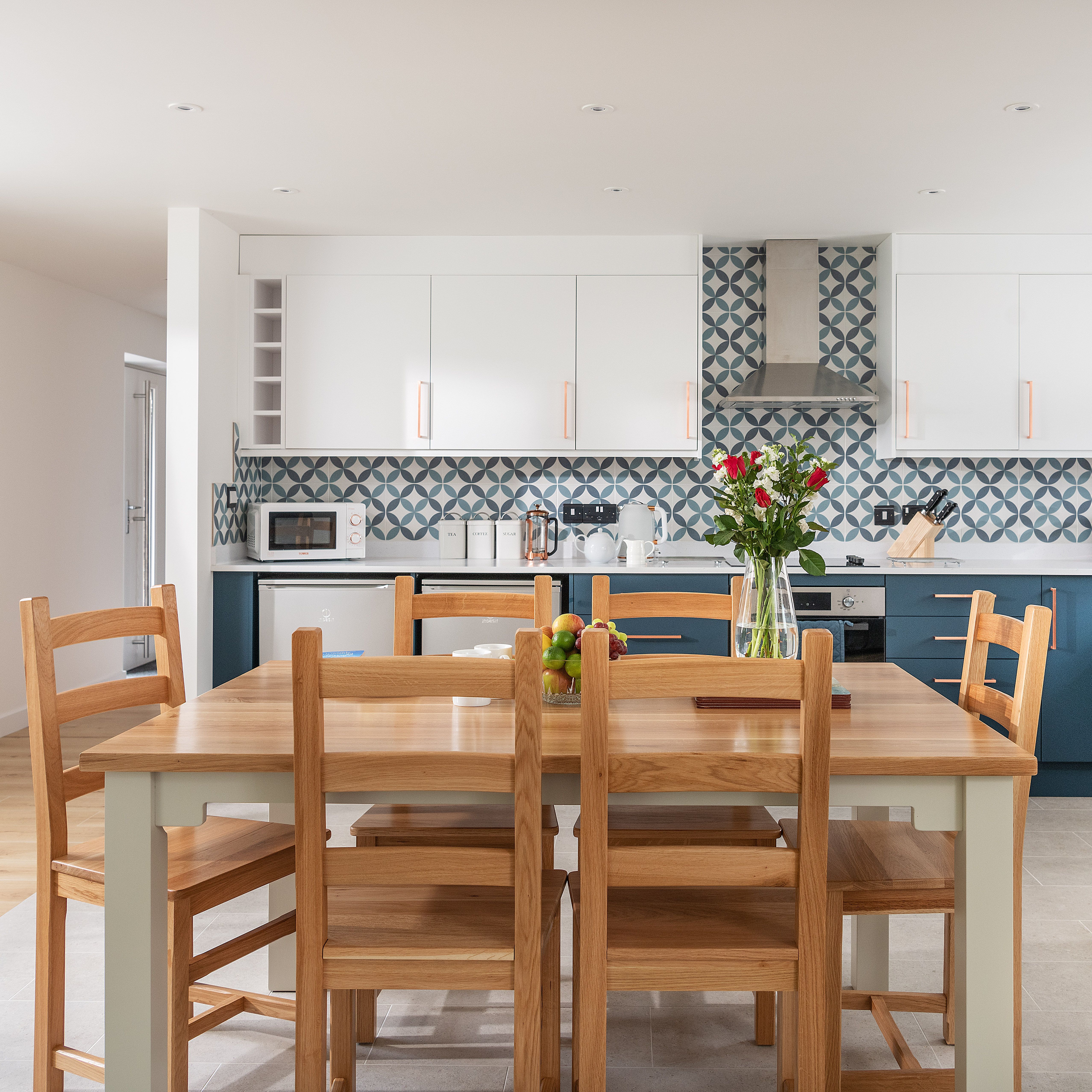 Modern kitchen with blue and white cabinets, patterned backsplash, wooden dining table with six chairs, vase of flowers and a bowl of fruit on the table, and stainless steel appliances.
