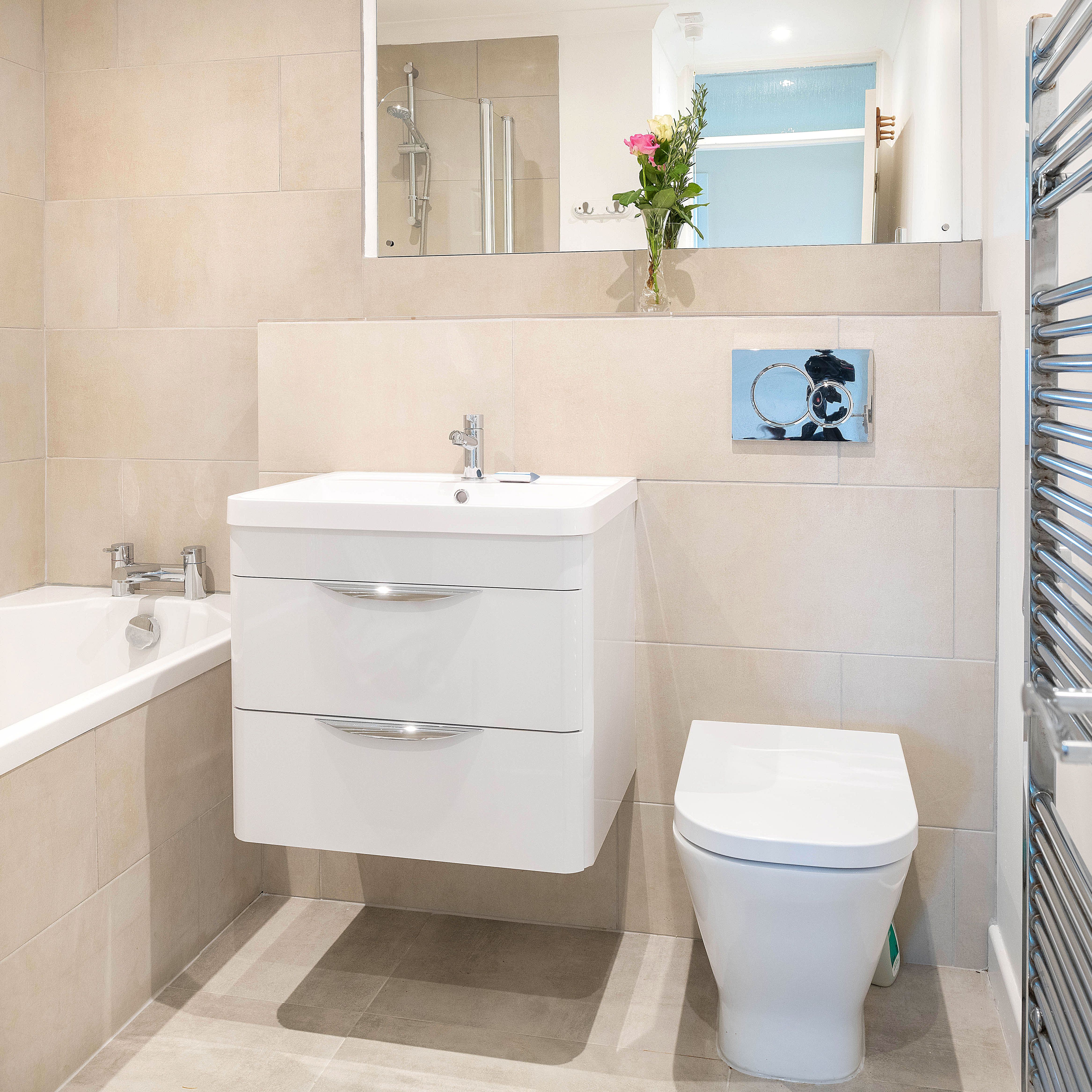 Modern bathroom with beige tiles, floating white vanity, toilet, bathtub, wall mirror, towel warmer, and a vase of flowers on the shelf.