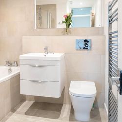 Modern bathroom with beige tiles, floating white vanity, toilet, bathtub, wall mirror, towel warmer, and a vase of flowers on the shelf.