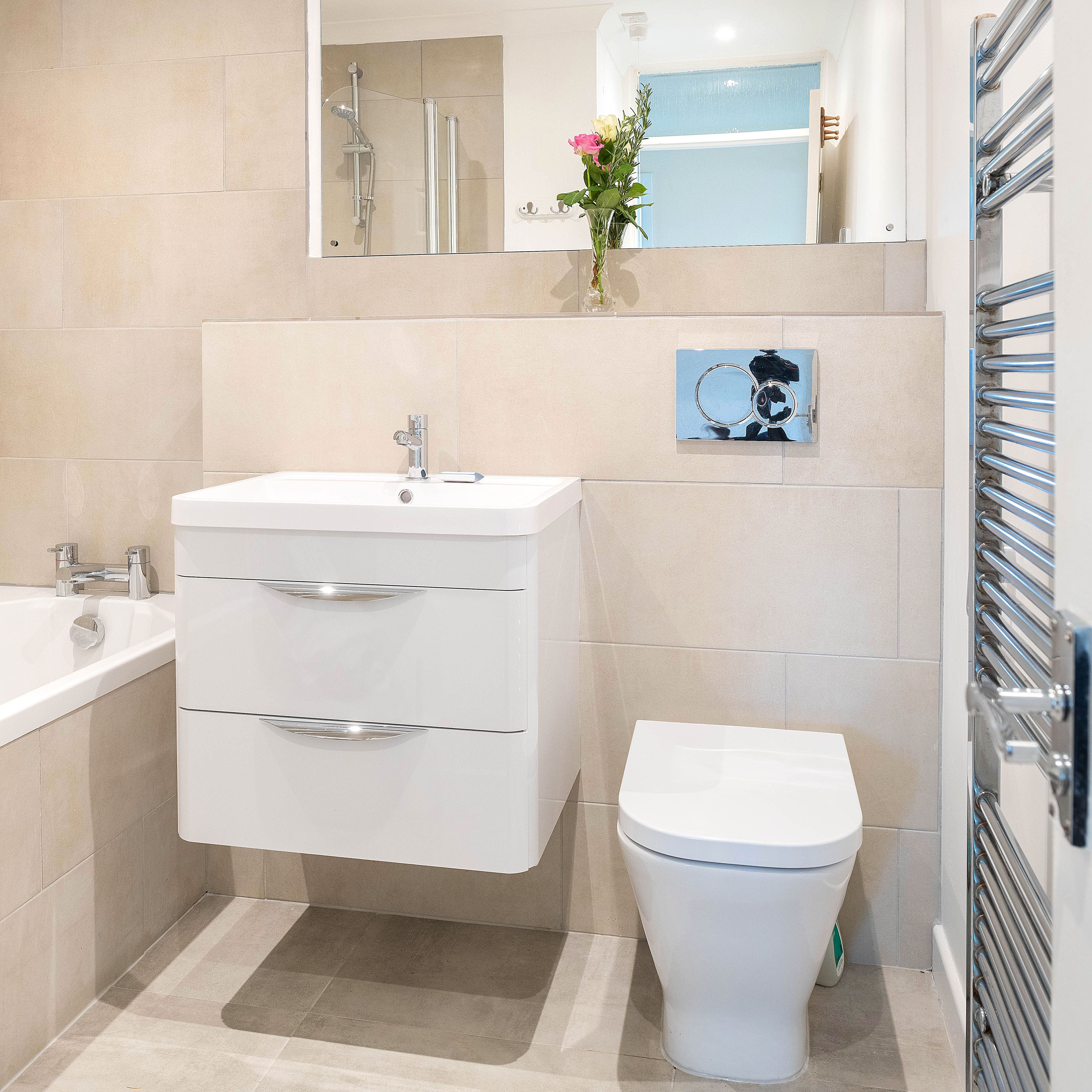 Modern bathroom with beige tiles, floating white vanity, toilet, bathtub, wall mirror, towel warmer, and a vase of flowers on the shelf.