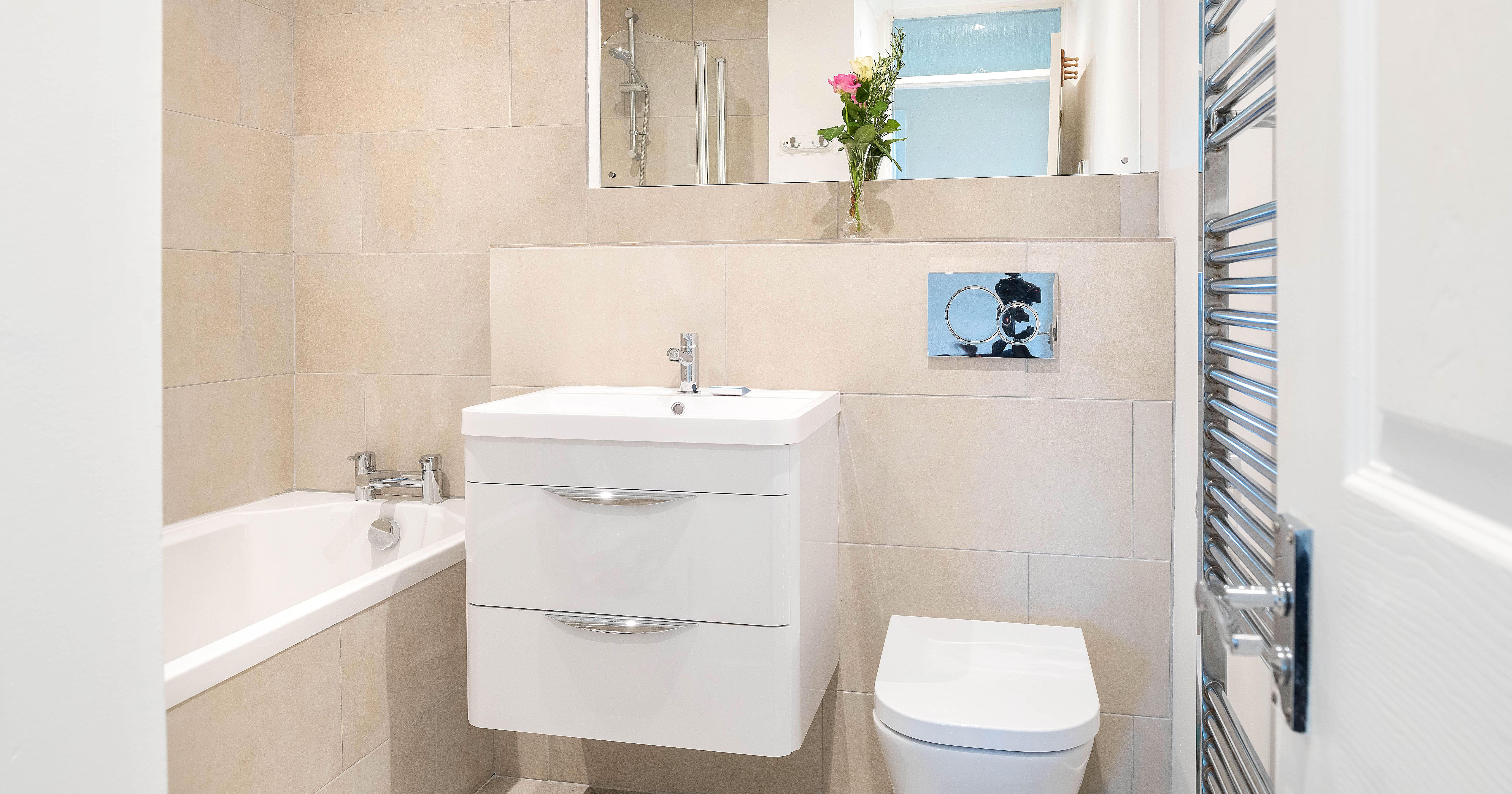 Modern bathroom with beige tiles, floating white vanity, toilet, bathtub, wall mirror, towel warmer, and a vase of flowers on the shelf.