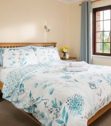 Bright and cozy bedroom with a wooden bed, floral bedding, bedside tables, a large window with blue curtains, and a chair with clothes draped over it.